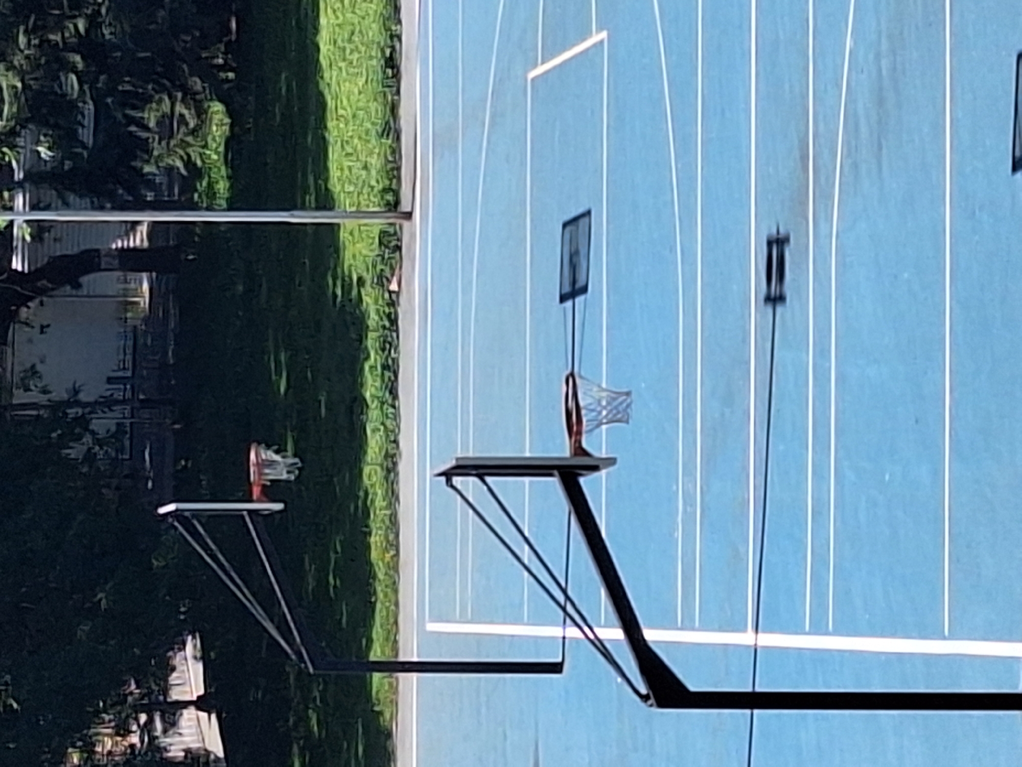 The image depicts an outdoor basketball court with a blue surface and white court lines. Two basketball hoops with orange rims and white nets are visible. One hoop is in the foreground, and another is in the background. Shadows of the hoops are cast on the court. Trees and buildings are visible in the background, suggesting an urban or suburban setting. The lighting indicates it is daytime, likely sunny. The scene appears to be in a public park or recreational area. The image does not show any people or specific landmarks that would allow for a precise identification of its location within Varna, Bulgaria.