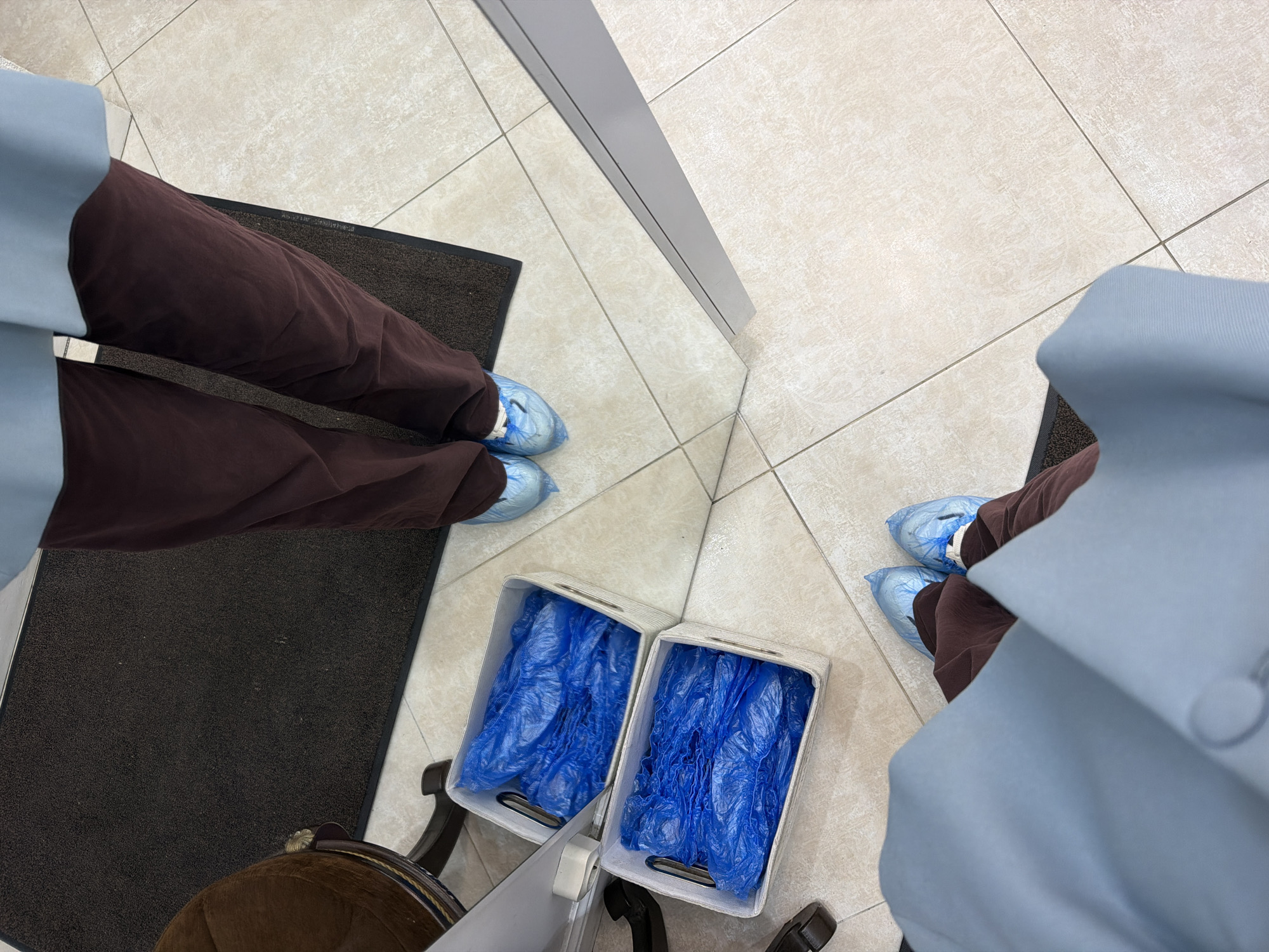 The photograph appears to be a selfie looking down at the feet of a person standing in front of a mirror. The person is wearing brown pants and light blue disposable shoe covers. Their clothing suggests a healthcare or laboratory setting. The mirror reflects the person and their surroundings. The floor is tiled in a neutral color. A black mat is visible near the person's feet. Next to the person's chair are two white containers filled with crumpled blue plastic bags. The location is not discernible. The city of Moscow, Russia is not able to be confirmed.