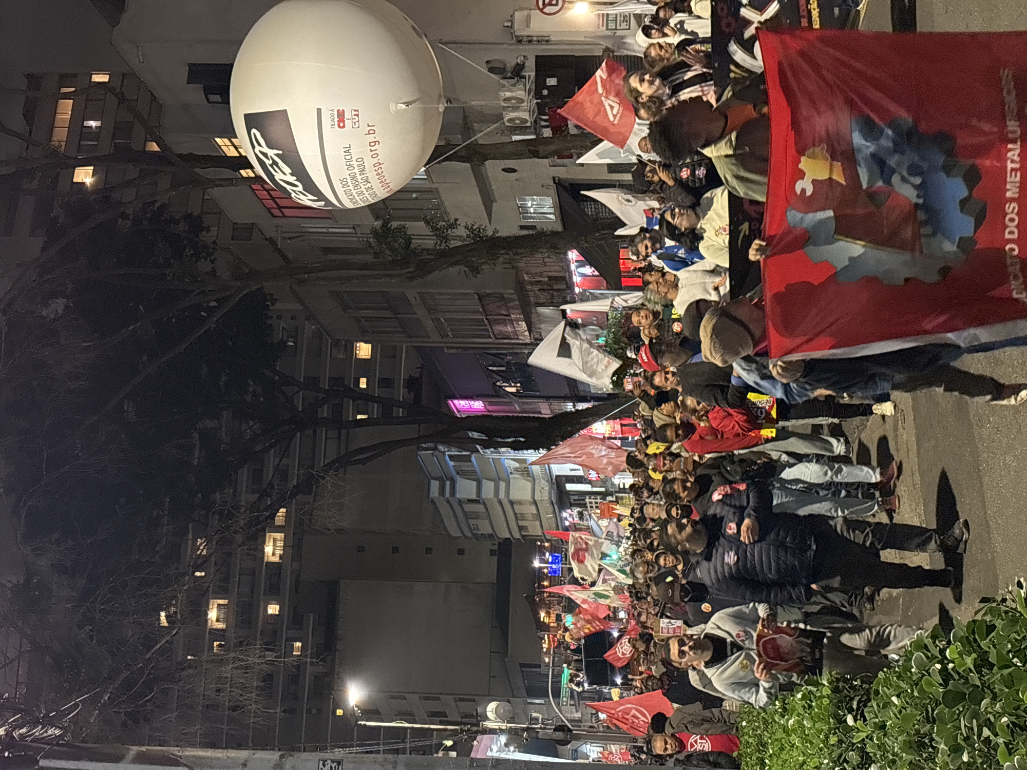 A medium-shot, full outdoor scene depicts a crowded street demonstration in São Paulo, Brazil, at night. The perspective is along the street, showing numerous people marching in a dense mass, holding red and white flags. One prominent red flag reads "SINDICATO DOS METALURGICOS" (Metallurgical Workers' Union).
Overhead, a large white balloon bears the message "APEOESP," an association of public school teachers in São Paulo. The website address "apeoesp.org.br" is also visible on the balloon, along with the acronyms "CINE" and "CUT". Buildings flank the street, some illuminated with lights on in various windows. The street is also lit by streetlights, contributing to the overall nighttime illumination. Trees are interspersed among the buildings, adding vertical elements to the scene. The foreground includes some green foliage, which serves as a visual barrier. The photo is taken from a slightly elevated angle. The image is rotated 90 degrees clockwise.