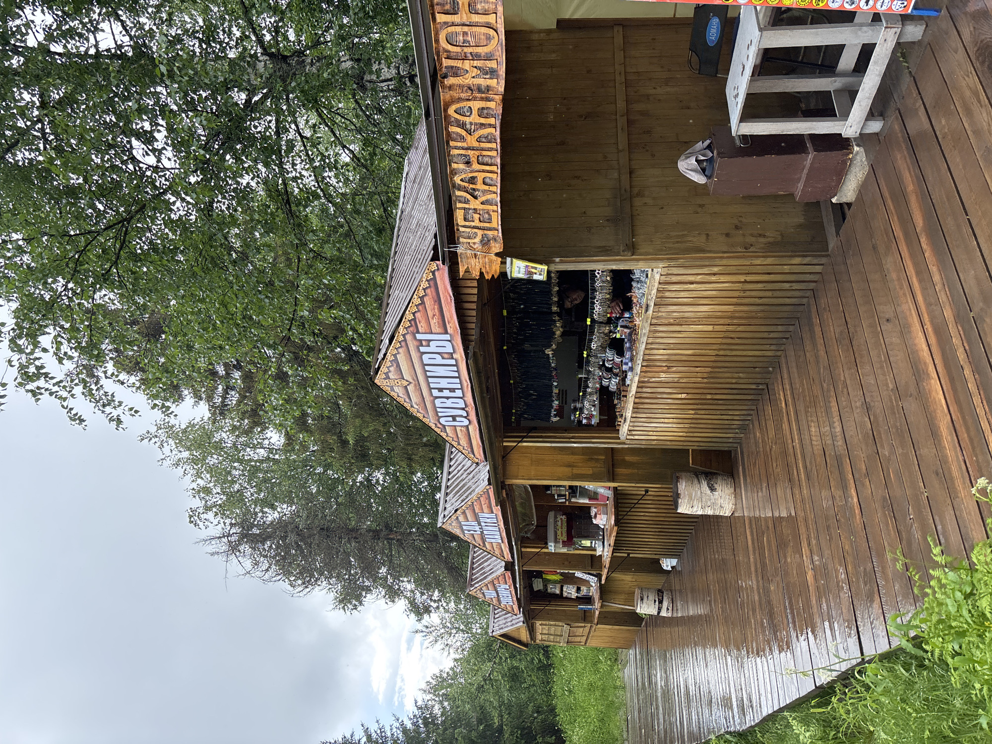 This is a vertical, eye-level, outdoor photograph of a row of souvenir kiosks on a wooden boardwalk. The boardwalk is wet and reflects the overcast sky. The kiosks are made of wood and have peaked roofs with ornate wooden trim. The kiosks are selling various items, including necklaces, bracelets, and what appear to be food and drinks. The signs above the kiosks are in Russian. The kiosk on the far right has a sign that reads "ЧЕКАНКА МОН" which translates to "minting coins". The kiosk to its left has a sign that reads "СУВЕНИРЫ" which translates to "souvenirs". There are trees behind the kiosks. The overall scene suggests a market or tourist area in a natural setting, likely near a body of water given the wet boardwalk. The image was captured in the city of Usvya, Russia.