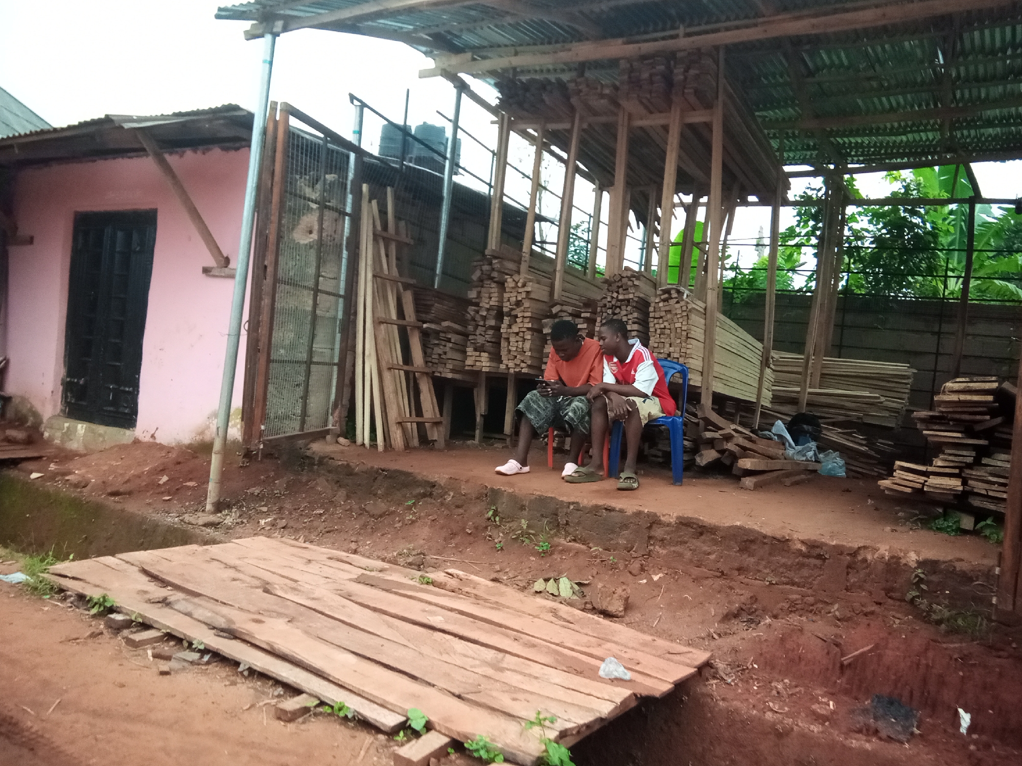 Two young males are seated on chairs outside a building, with stacks of lumber visible in the background. The male on the left is wearing a red shirt and patterned shorts. He is looking down at his phone. The male on the right is wearing a red and white striped shirt and patterned shorts, and he is looking to his left. The building has a pink exterior and a dark-colored door. A metal gate is visible next to the door. The ground is composed of red soil, and there is a wooden structure covered with a corrugated metal roof behind the seated individuals. The area appears to be a lumber yard or workshop. This scene is located in Oko Town, Nigeria.