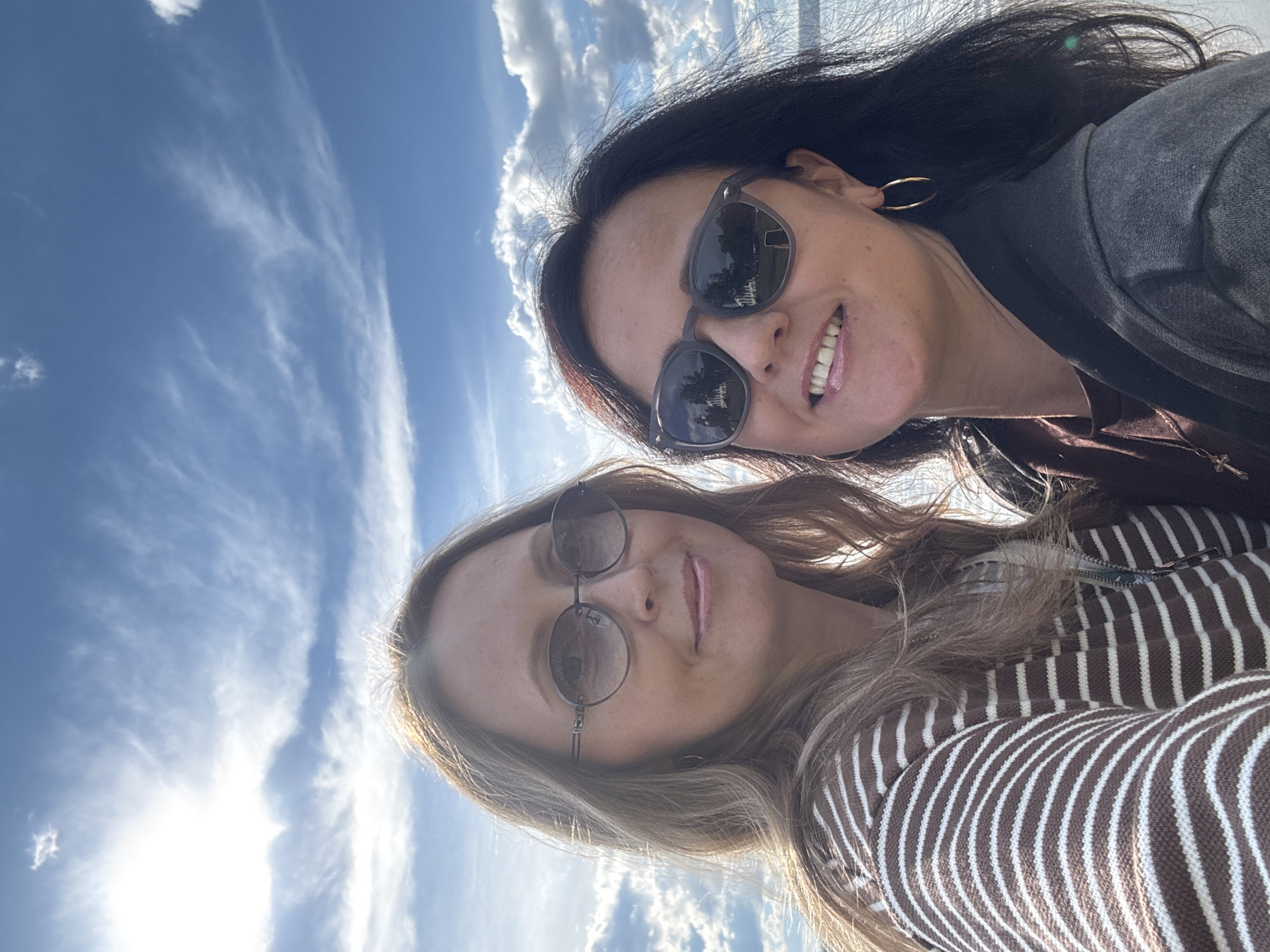 Two women are visible in a selfie. The woman at the top has dark hair, sunglasses, and is wearing a dark jacket over a brown shirt, with hoop earrings. The woman at the bottom has light brown hair, sunglasses, and is wearing a brown and white striped shirt. The background shows a blue sky with white clouds. The location is not identifiable as Votkinsk, Russia.