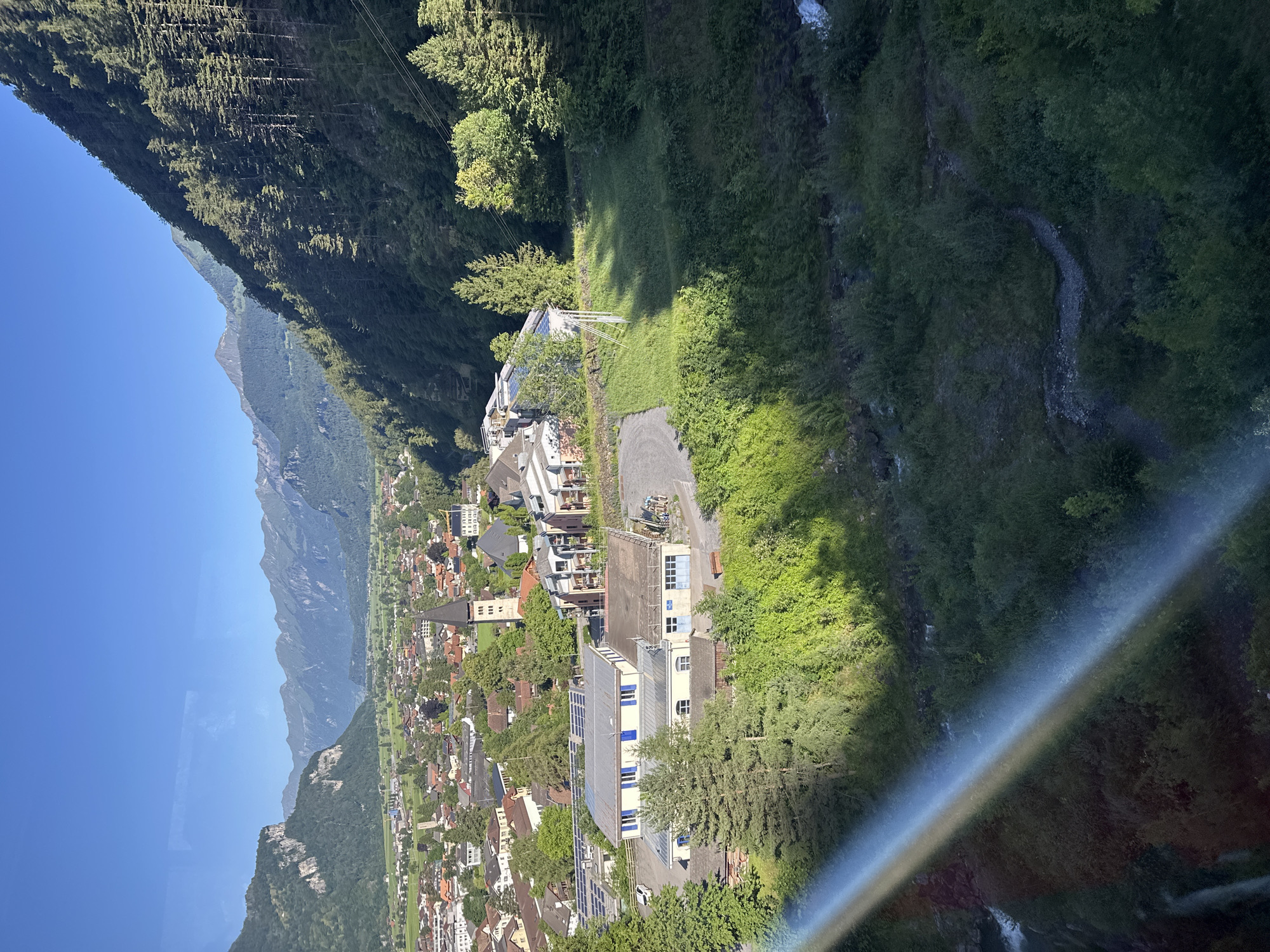 This is a high-angle, wide-shot, landscape photograph of the village of Hüsen, Switzerland, taken on a sunny day. The foreground is dominated by a lush, green, forested mountainside and a small stream. A rainbow-like glare streaks across the lower left portion of the image. In the midground, several buildings are visible, including a light-colored, rectangular building with a silver roof covered in solar panels. A church with a dark steeple is visible in the distance. The background features a wide valley with a small town nestled within it, surrounded by green fields and imposing, rocky mountains. The sky is clear and blue. No people are visible in the photograph.