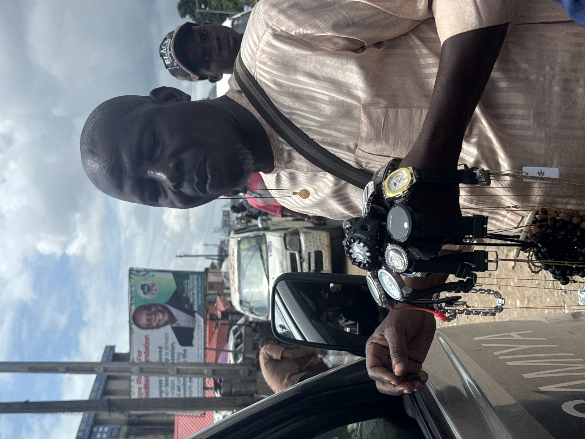 The media file shows a man selling watches and necklaces from a car. He is wearing a light-colored, striped shirt and has several watches on his right arm. Behind him, another man is visible wearing a "BLACK" cap. The scene appears to be outdoors, possibly a street, with a billboard featuring a man's smiling face and text in the background. The sky is overcast with clouds, suggesting it might be daytime. The setting includes traffic and other vehicles, indicating a busy urban environment. The man selling watches seems to be interacting with potential customers from his car. Some of the visible text includes "SAMINA" on the car's surface and partial text on the billboard that appears to be in English.