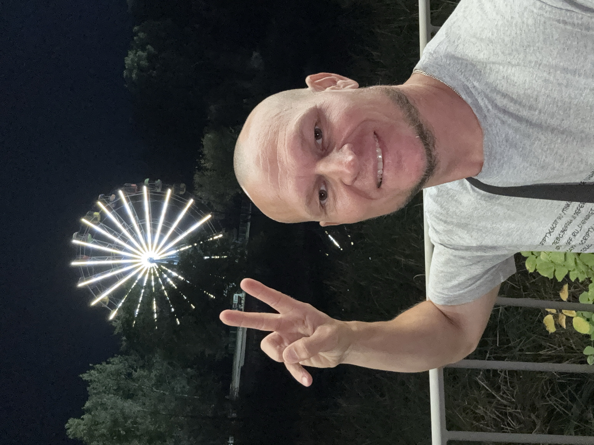 A man with a shaved head and a beard smiles at the camera, holding up two fingers in a peace sign. He is wearing a gray t-shirt. In the background, a Ferris wheel is lit up at night. The scene appears to be an amusement park or fairground. The location is the city of Oskemen, Kazakhstan.