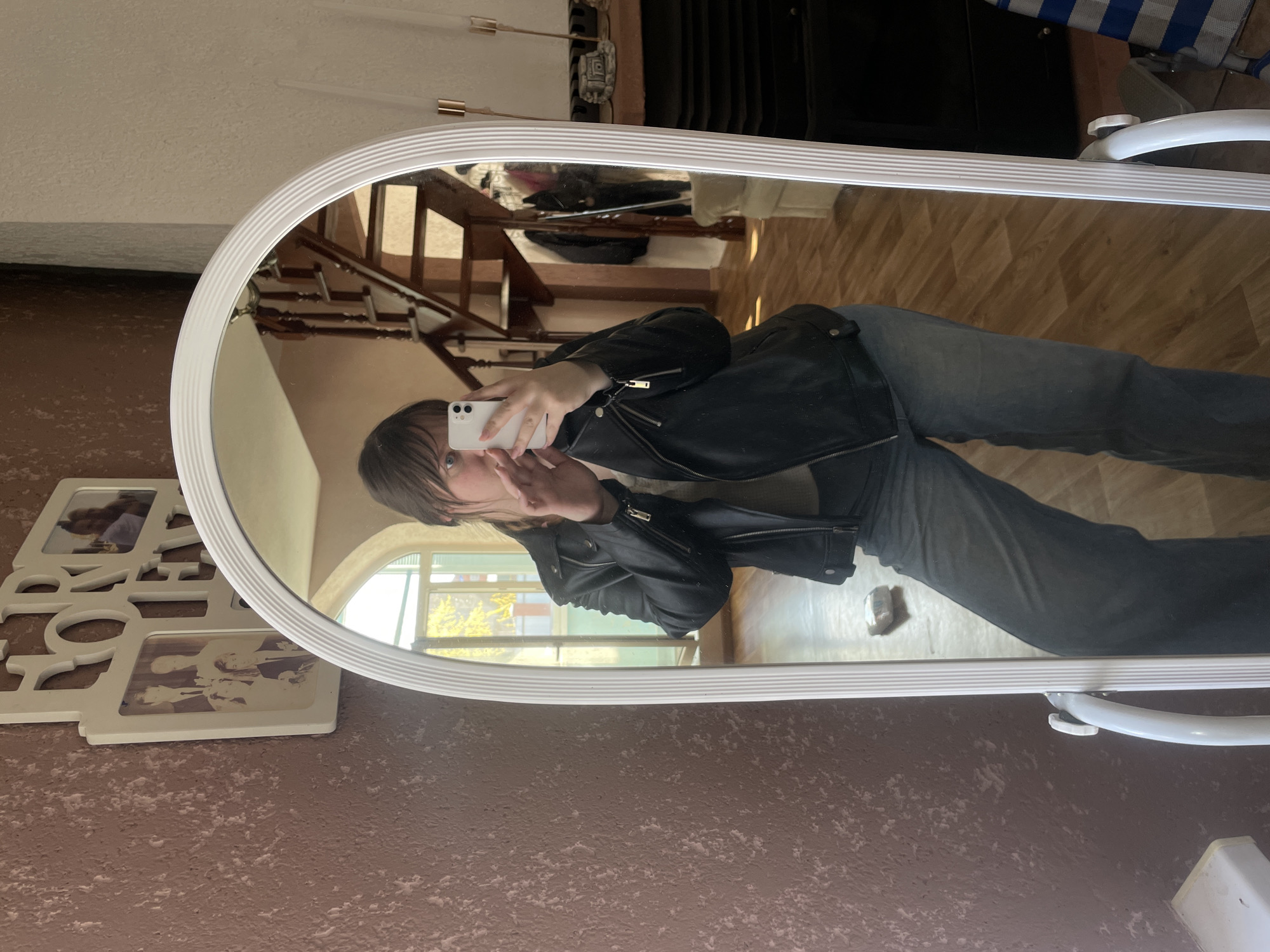 A person reflected in a white-framed mirror is holding a white cell phone and taking a selfie. The person is wearing a black leather jacket and jeans. The reflection also shows a staircase, a window, and part of the room. A framed picture saying "Home" is on the wall to the left of the mirror.
The location is not specified as the city of Noviy, Russia.