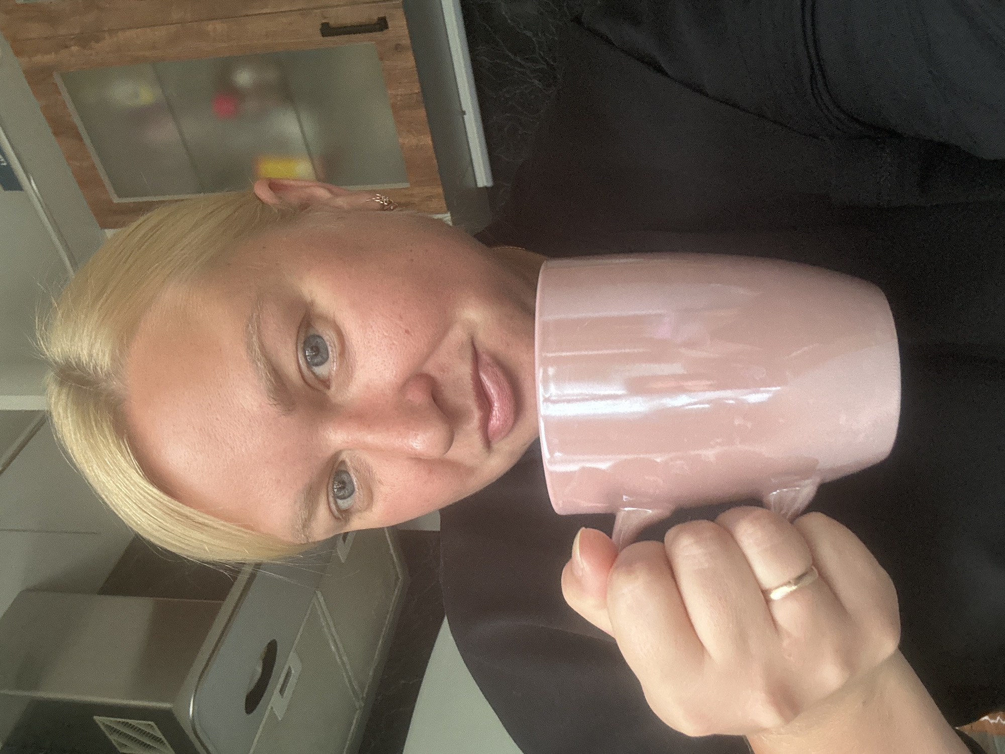 A medium shot captures a woman with blonde hair and light blue eyes holding a pink mug with her right hand. She is wearing a black shirt. A silver wedding band is visible on her left ring finger. In the background, a kitchen environment is suggested by cabinetry and appliances. The image is taken from a slightly elevated angle, looking down at the woman. The scene is indoors, likely a residential kitchen. The lighting is soft, and the overall tone is casual. The city of Chaikovsky, Russia, is not discernible from the image.