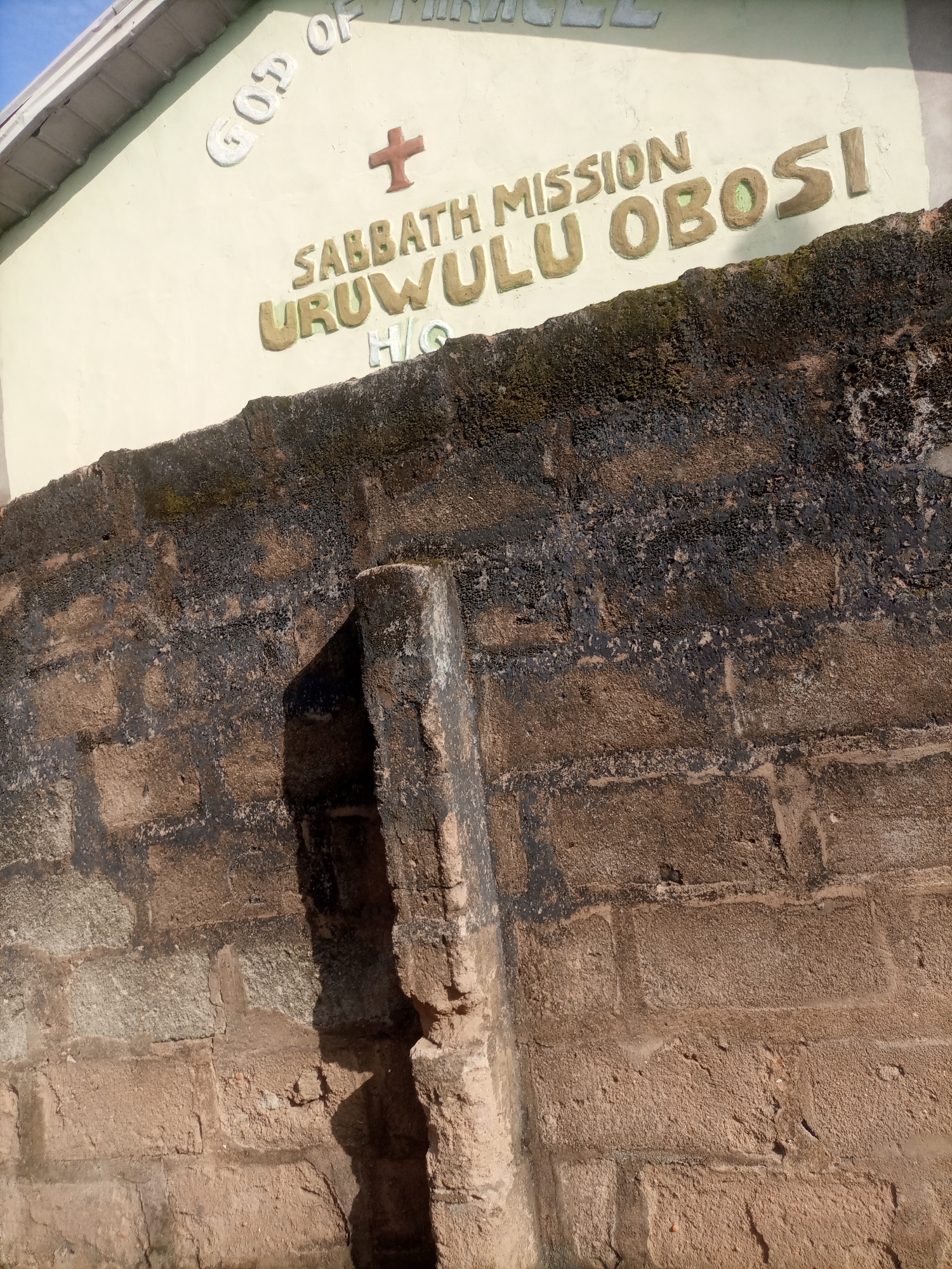 The image shows the exterior of a building in Nkpor, Nigeria, with "GOD OF MIRACLE SABBATH MISSION URUWULU OBOSI H/C" inscribed in raised lettering on its cream-colored facade. A red cross symbol is also visible on the wall. The building is partially obscured by a weathered, dark brown brick wall in the foreground, which shows signs of moss and age. The wall is constructed of rough-hewn stones and bricks, some of which are darker and appear to be stained. The overall scene is outdoors, under a bright sky, suggesting daytime.