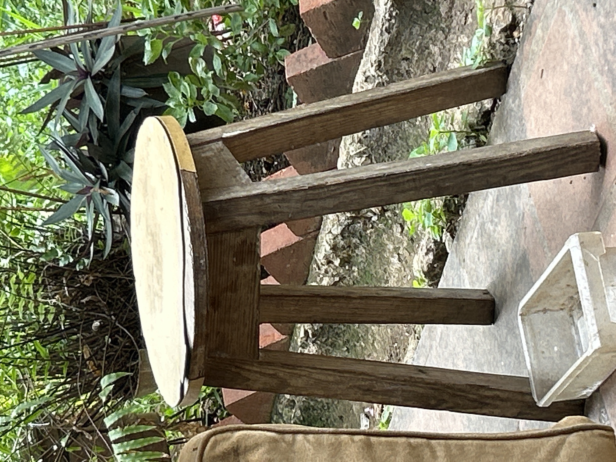 The image shows a weathered wooden stool with a round, light-colored top, standing on a concrete surface. The stool has four legs and is situated outdoors, surrounded by dense green foliage. In the background, there are some reddish-brown bricks arranged in a staggered pattern along a wall. To the left of the stool, a portion of a tan-colored upholstered object, possibly a chair or cushion, is visible. In front of the stool, on the concrete, is a clear plastic container with some dirt or debris inside. The environment appears to be a garden or courtyard, with plants growing in pots and along the ground. The lighting suggests it is daytime. The specific location in Santa Ana, Venezuela, cannot be determined from the visual information provided in the image.
