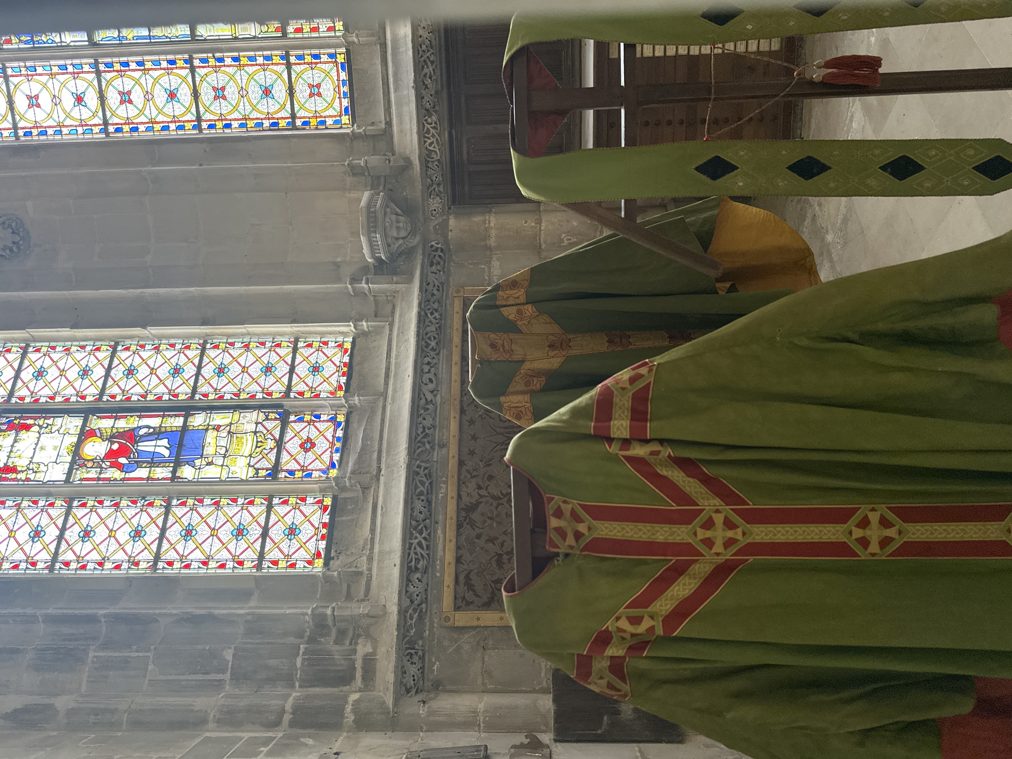 This image depicts the interior of a church, likely in Dieppe, France, featuring religious vestments displayed in the foreground. Three green chasubles, ornate liturgical garments, are visible. The chasuble in the immediate foreground is predominantly green with red and gold decorative bands featuring crosses and intricate woven patterns. To its right, another green chasuble is draped, revealing a gold-lined interior. A third vestment, a green stole with a diamond pattern in black and red tassels, hangs on the right side of the frame. In the background, large stained-glass windows with colorful geometric patterns and religious figures are illuminated by natural light, casting vibrant hues onto the stone walls. Architectural details such as carved stone friezes and columns are also present. The scene is static, with no people or discernible actions. The lighting suggests an overcast or indoor setting, with the stained glass being a primary light source.