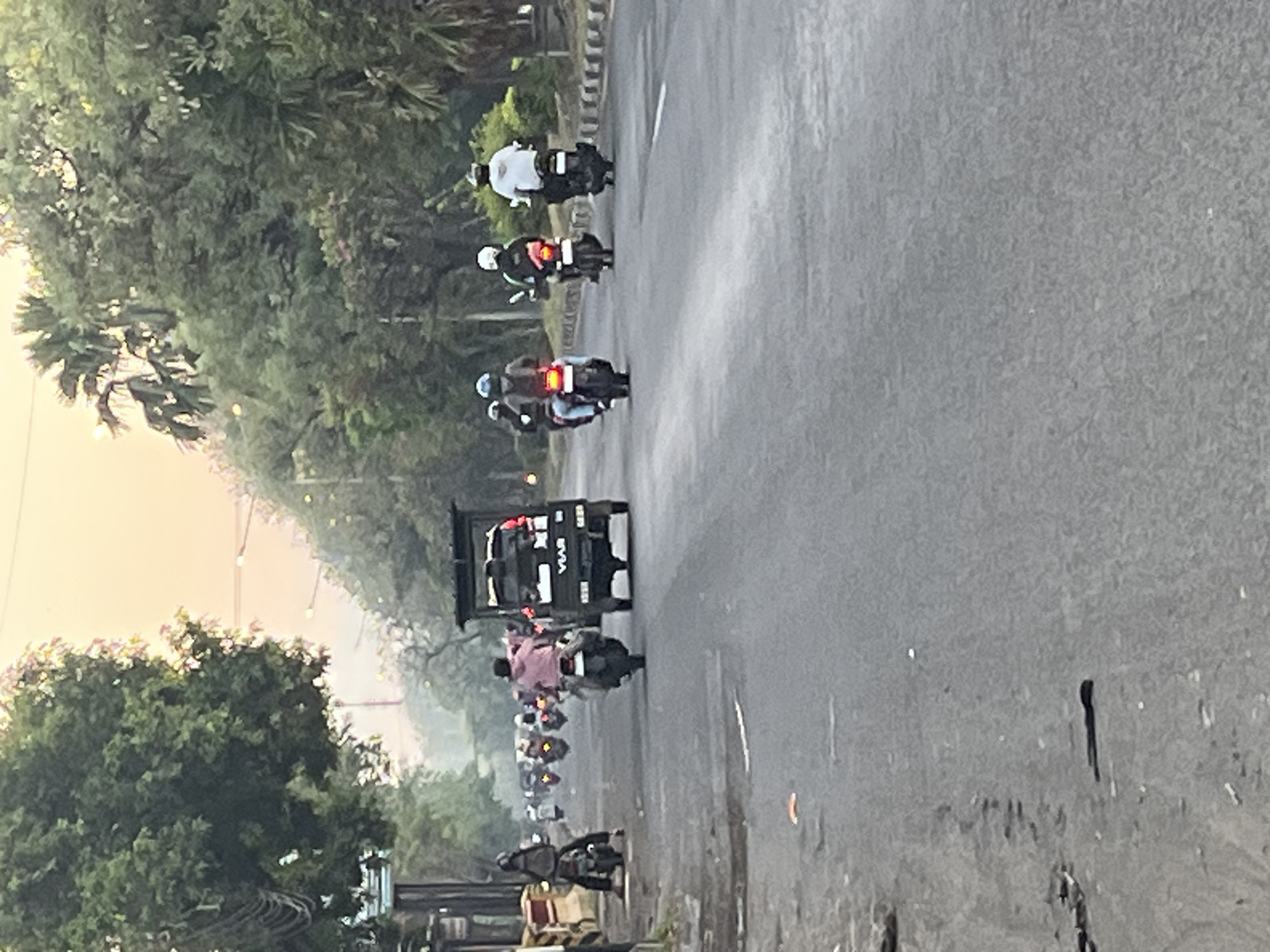 A slightly blurry, eye-level medium shot shows a street scene at what appears to be dawn or dusk in Ngaban, Indonesia. The sky is a soft, hazy orange and pink, suggesting early morning or late afternoon light. Lush green trees line the sides of the road, with some palm trees visible further in the background. The road itself is asphalt and appears to be a two-lane street.
Several motorcycles and a small truck are present. In the foreground, on the left side of the road, a person on a motorcycle is parked or moving slowly. Further down the road, a black truck with "VIAR" branding on the back is driving away from the camera, carrying some cargo. Behind the truck, a line of motorcycles are also traveling in the same direction. One motorcycle has a passenger wearing a pink jacket and a black helmet. Other motorcycles have riders wearing helmets, some with light-colored jackets. The motorcycles are spaced out, indicating normal traffic flow. The road surface shows some signs of wear, with patches and debris. The overall impression is a typical urban street scene during a transitional time of day.