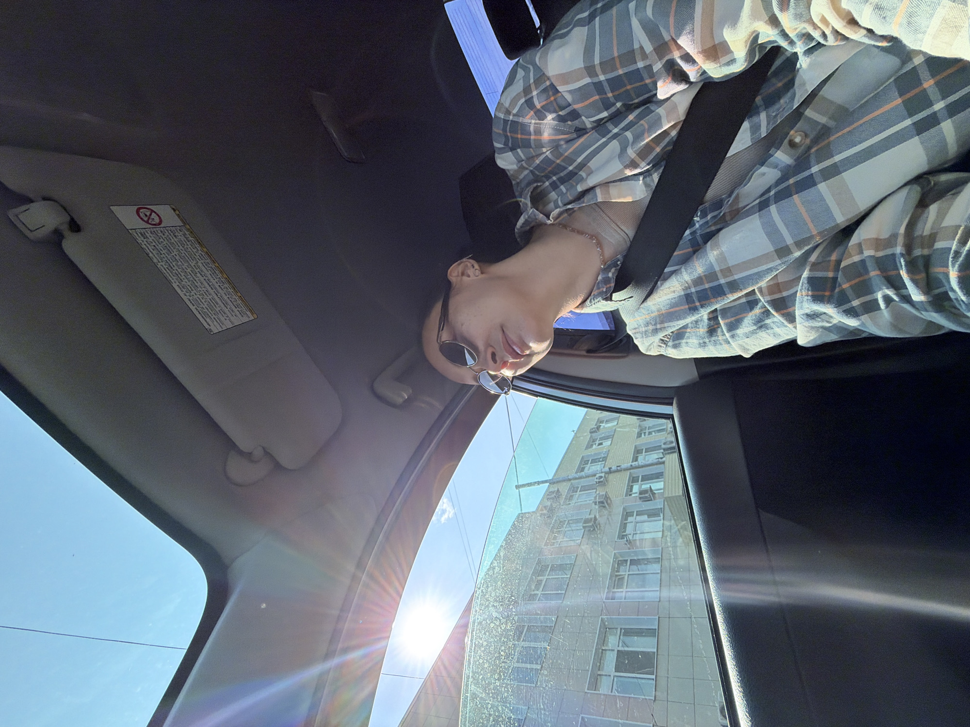 A woman wearing sunglasses and a plaid shirt is in the passenger seat of a car. She is looking out the window at a sunlit building. The sun is shining brightly, casting lens flare across the image. The car interior is visible, including the dashboard and a sun visor with a warning sticker. The scene appears to be in the city of Chaikovsky, Russia, based on the architecture of the building.