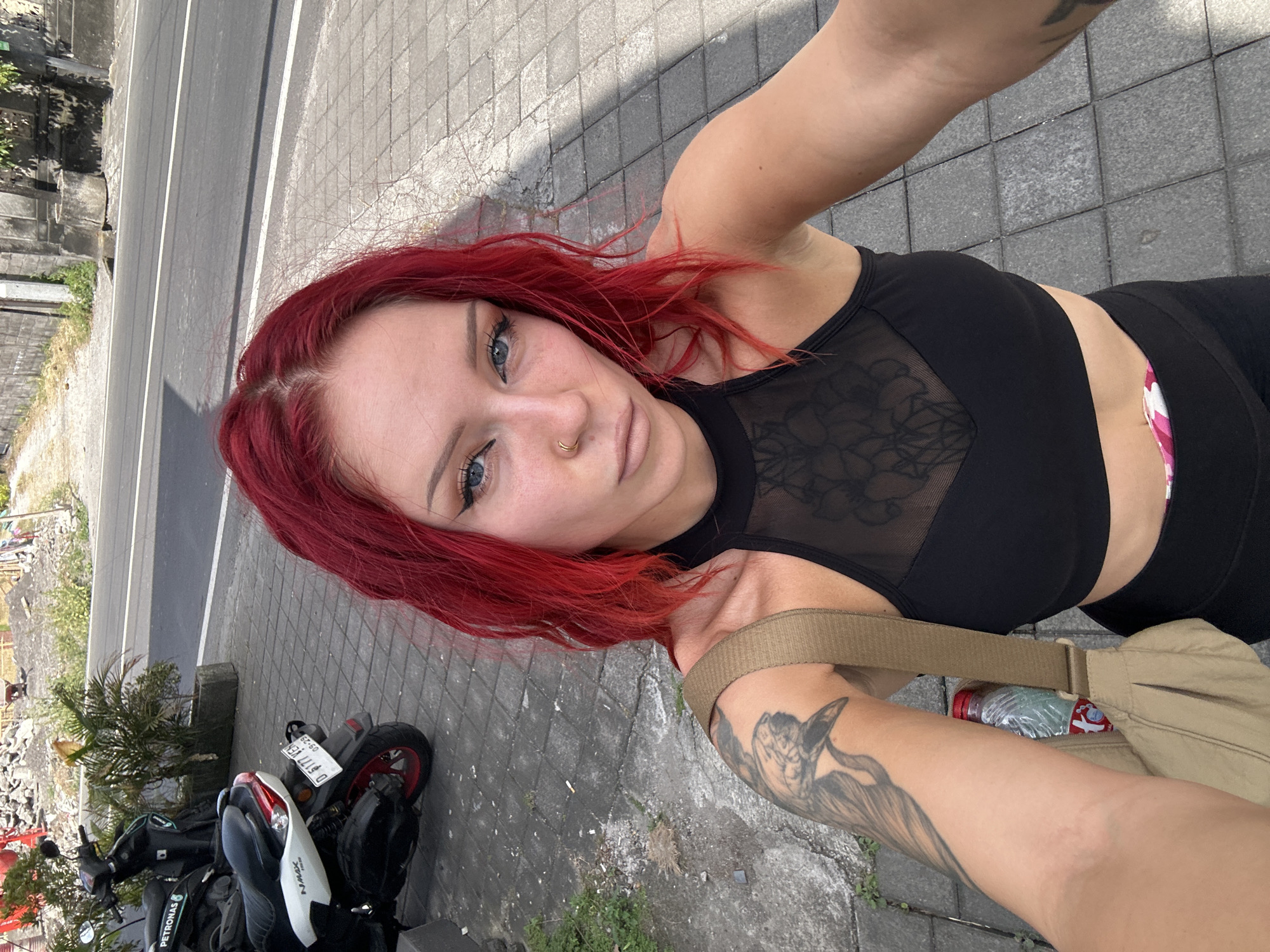 A woman with bright red hair and blue eyes, wearing a black crop top and black leggings, stands on a paved sidewalk in North Kuta, Indonesia. She is taking a selfie with her right arm extended. A tan messenger bag with a water bottle inside is slung across her body. Two parked scooters are visible to her left, and a road with a white line runs parallel to the sidewalk. Construction debris and some vegetation are present in the background.