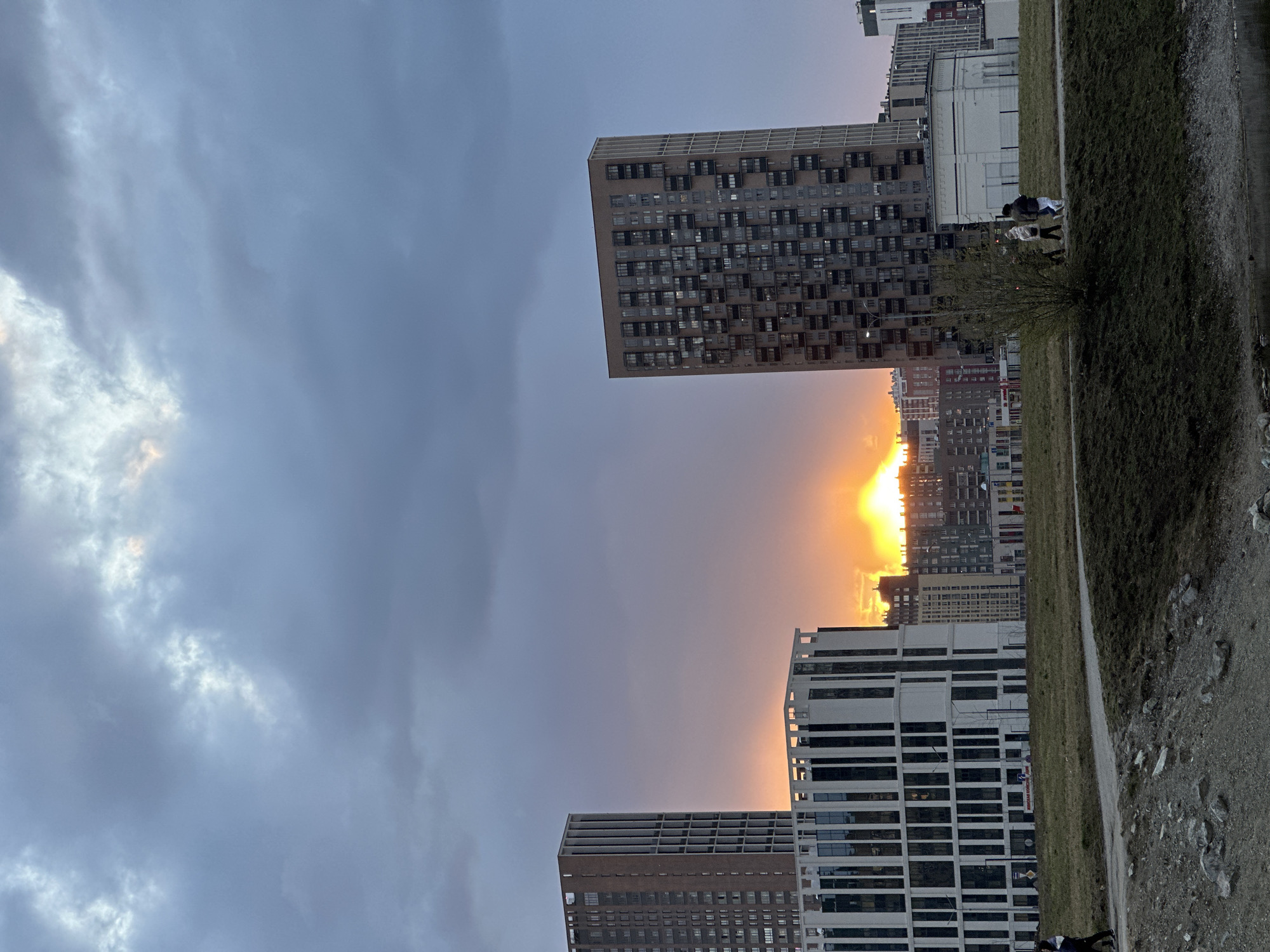 The photograph depicts a cityscape at sunset, with buildings and parkland visible in Yekaterinburg, Russia. The sky is cloudy, with the sun appearing between buildings in the distance, casting an orange glow. Two tall buildings stand out in the foreground. The scene includes a grassy park area. Two individuals are walking in the background. The image is oriented vertically.