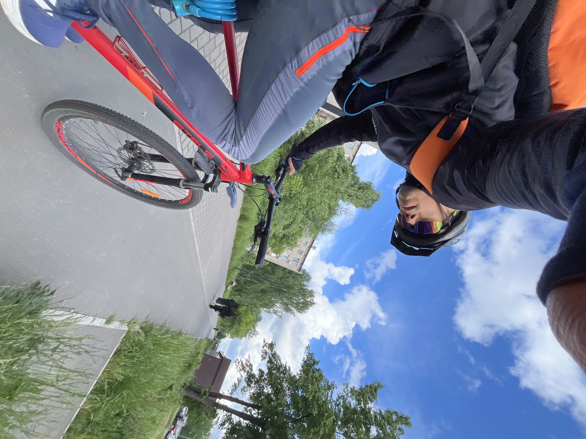 A person is captured in a low-angle selfie while riding or stopped on a red mountain bike in Chaikovsky, Russia. The cyclist wears a dark long-sleeved top, grey athletic pants with orange and light grey accents, a dark cycling helmet, and reflective sunglasses. A black backpack with orange shoulder straps is also visible. The photo is taken looking upwards, featuring the rider's face and upper body prominently against a bright blue sky with scattered white clouds.
The red bicycle has black and orange details, with the brand name "STELS" clearly visible on the frame. The front wheel features a knobby tire with the text "TOUGH TOM" visible on its sidewall, and it is equipped with disc brakes. A blue water bottle holder is attached to the bicycle's frame.
The setting appears to be an outdoor urban or suburban environment on a clear, sunny day. The cyclist is on a paved path or road, flanked by green grass and trees. In the background, multi-story residential buildings are visible, indicating a developed area. The overall impression is one of active recreation in pleasant weather.