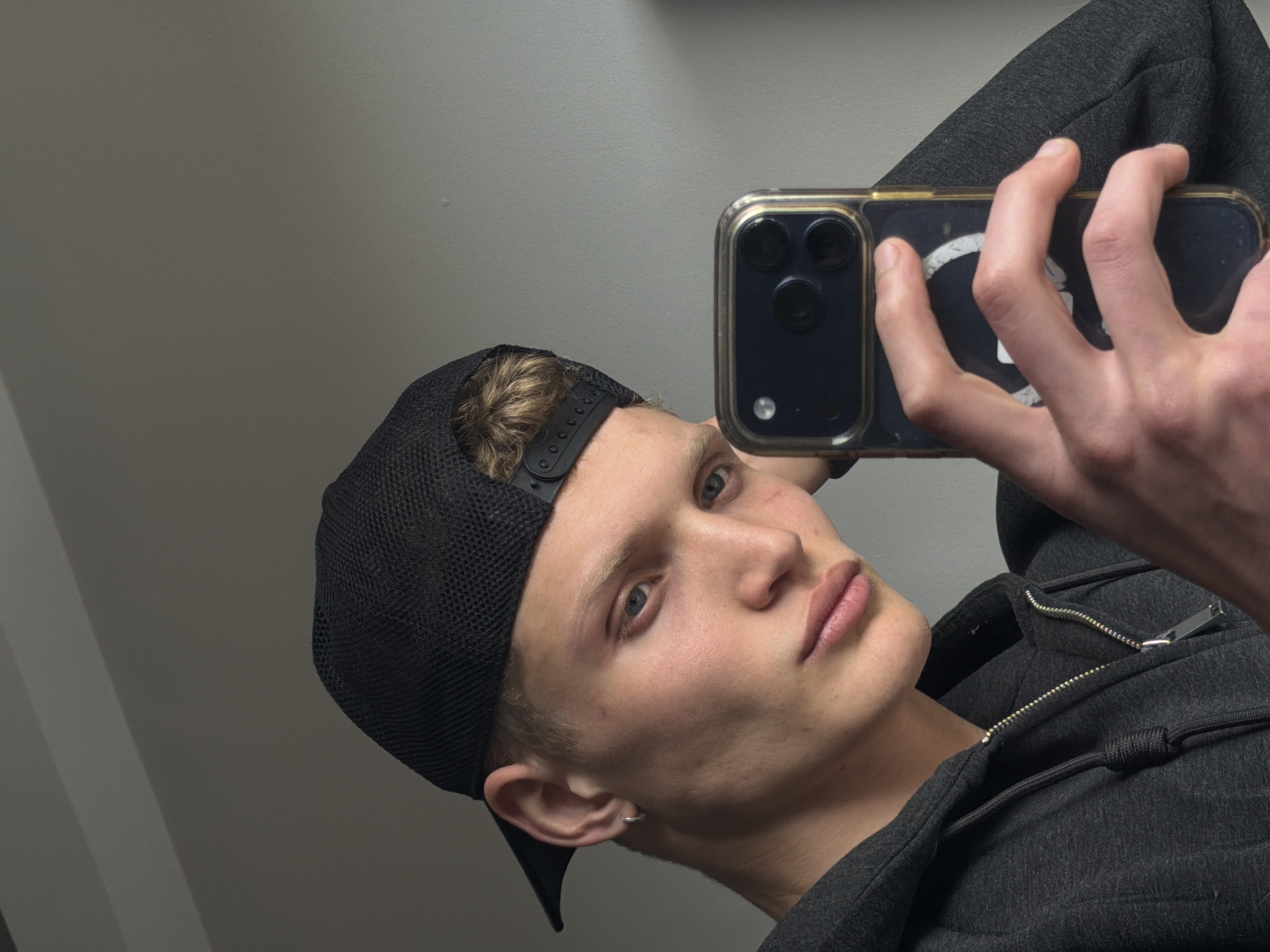 A young, light-skinned male with short blond hair and blue eyes is taking a selfie. He is wearing a black baseball cap and a zipped-up dark gray hoodie. He is holding a smartphone with a gold-trimmed case in his right hand, obscuring the camera's view. He has a small hoop earring in his left ear. The background is a plain light gray wall. The location cannot be determined as London, United Kingdom.
