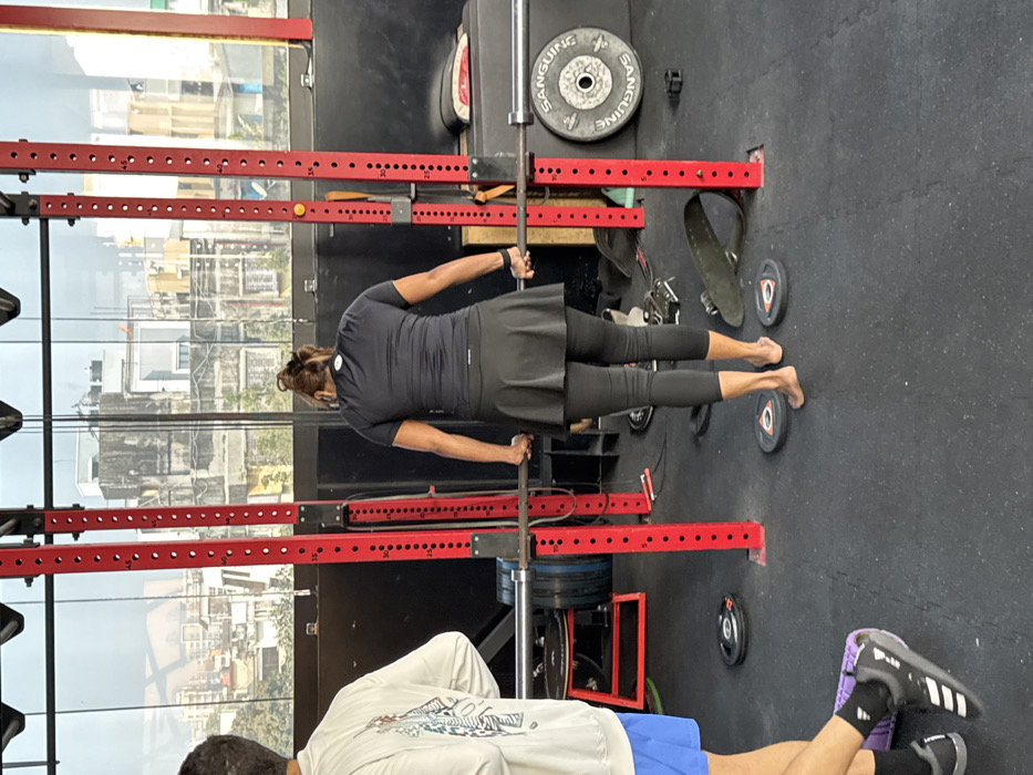 In a gym setting, a woman wearing black athletic attire is performing a gymnastic ring exercise. The rings are supported by a red squat rack, and weight plates are visible around the rack. A man's legs and feet are at the lower edge of the image. The background shows a cityscape through a window.
