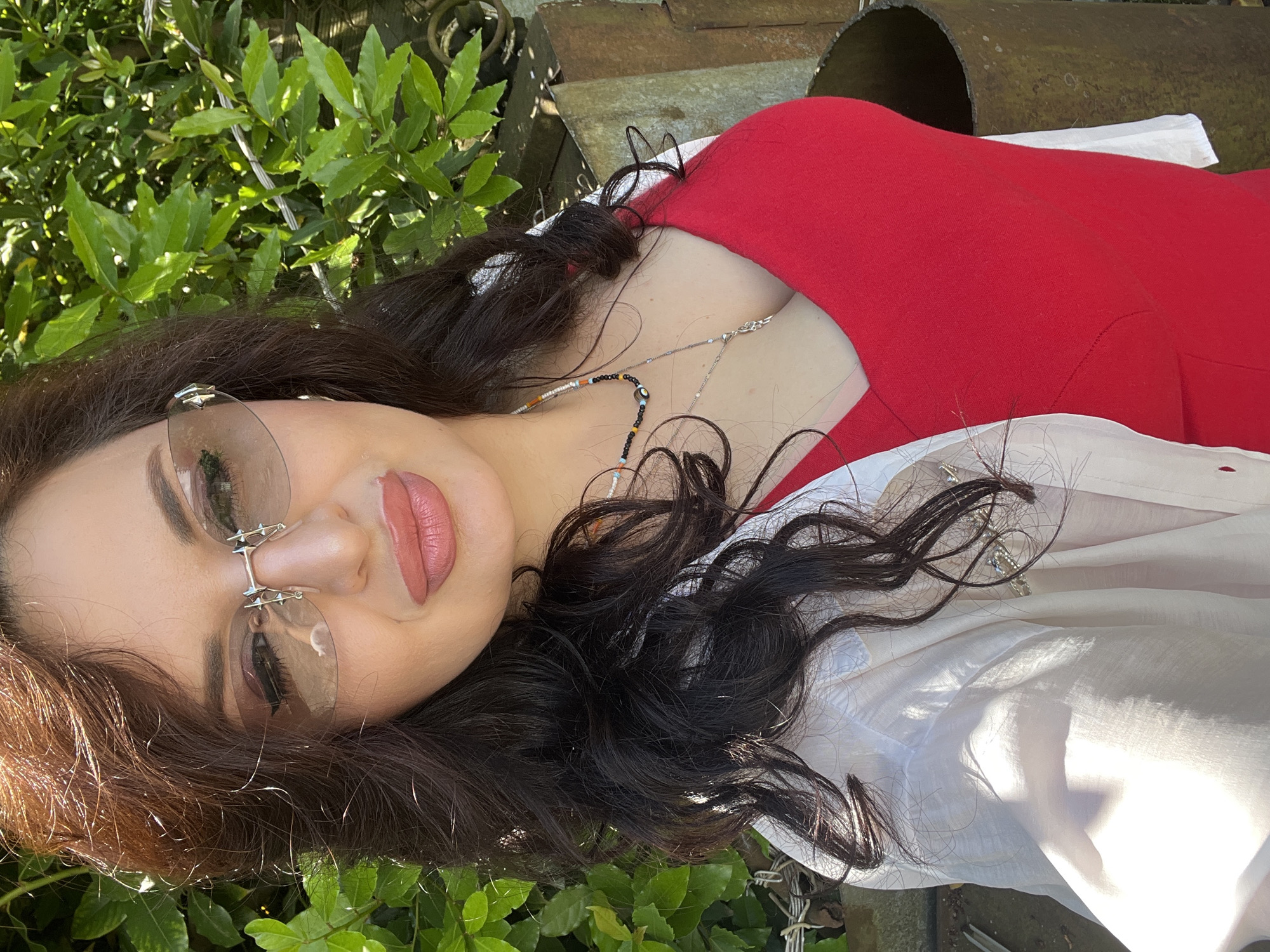 A woman is taking a selfie outdoors. She has long, dark, wavy hair and is wearing a red top and a white unbuttoned shirt over it. She has on unique silver-rimmed sunglasses with spikes on the bridge, and several necklaces, one of which has colorful beads and an evil eye charm. Her makeup includes winged eyeliner and pink lipstick. She is smiling slightly and her head is tilted. The background consists of lush green foliage and some weathered metal structures. The lighting suggests it is a sunny day.