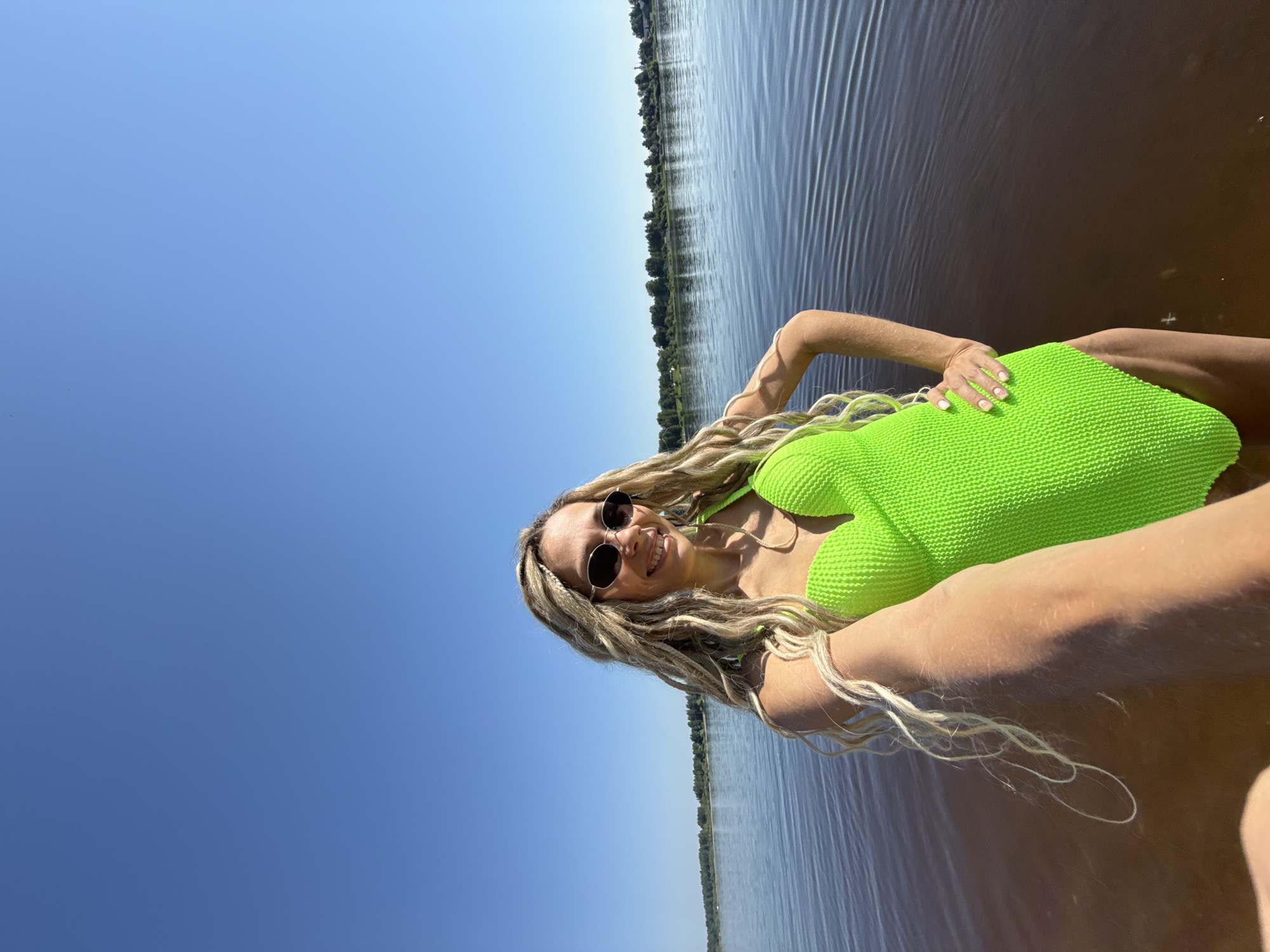 A medium shot captures a woman with blonde dreadlocks, wearing oval sunglasses and a vibrant green one-piece swimsuit, standing in shallow, murky water under a clear blue sky. She is facing the camera, holding the camera out to take a selfie with her right arm extended. Her left hand rests on her hip. The background features a body of water with ripples, a tree line on the distant shore, and a clear sky. The location is identified as Rusanovo, Russia.