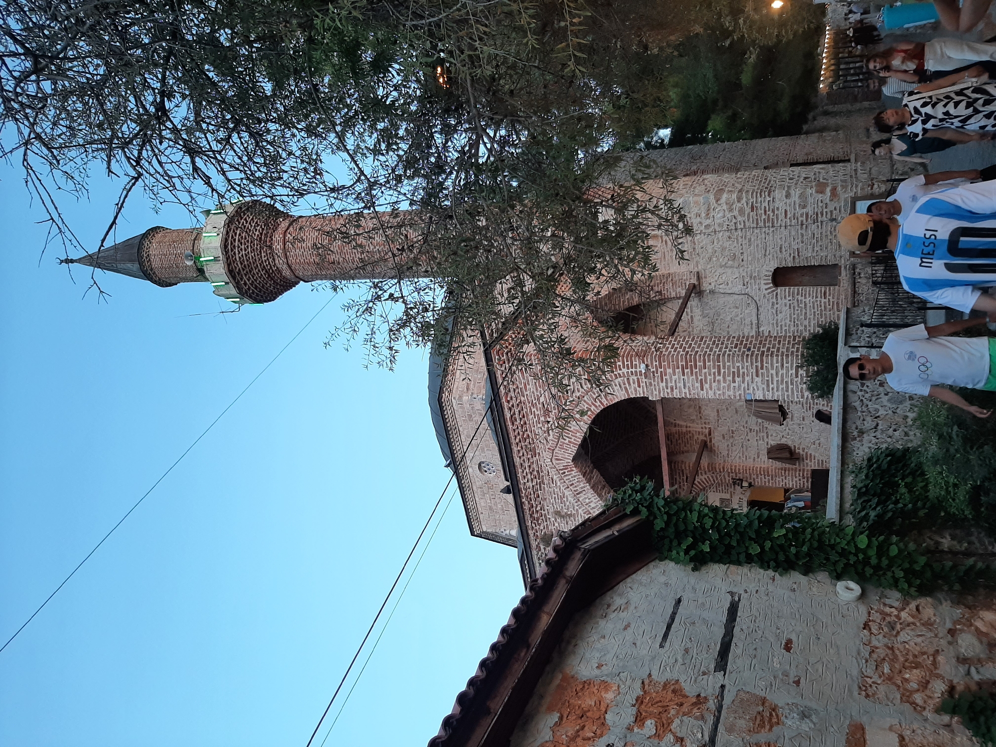 In Alanya, Turkey, a brick mosque with a tall, conical minaret stands prominently against a clear blue sky. The minaret features green lights near its top. In the foreground, people walk along a cobblestone path. A man in the foreground wears a white t-shirt with Olympic rings and green shorts. Behind him, another man is seen from the back wearing a blue and white soccer jersey with the name "MESSI" and the number "10" on it. To the left of the path, an old stone building is partially covered in green ivy. Trees with dark leaves partially obscure the view of the mosque and minaret. The overall scene suggests a street or plaza area adjacent to a historic religious site.