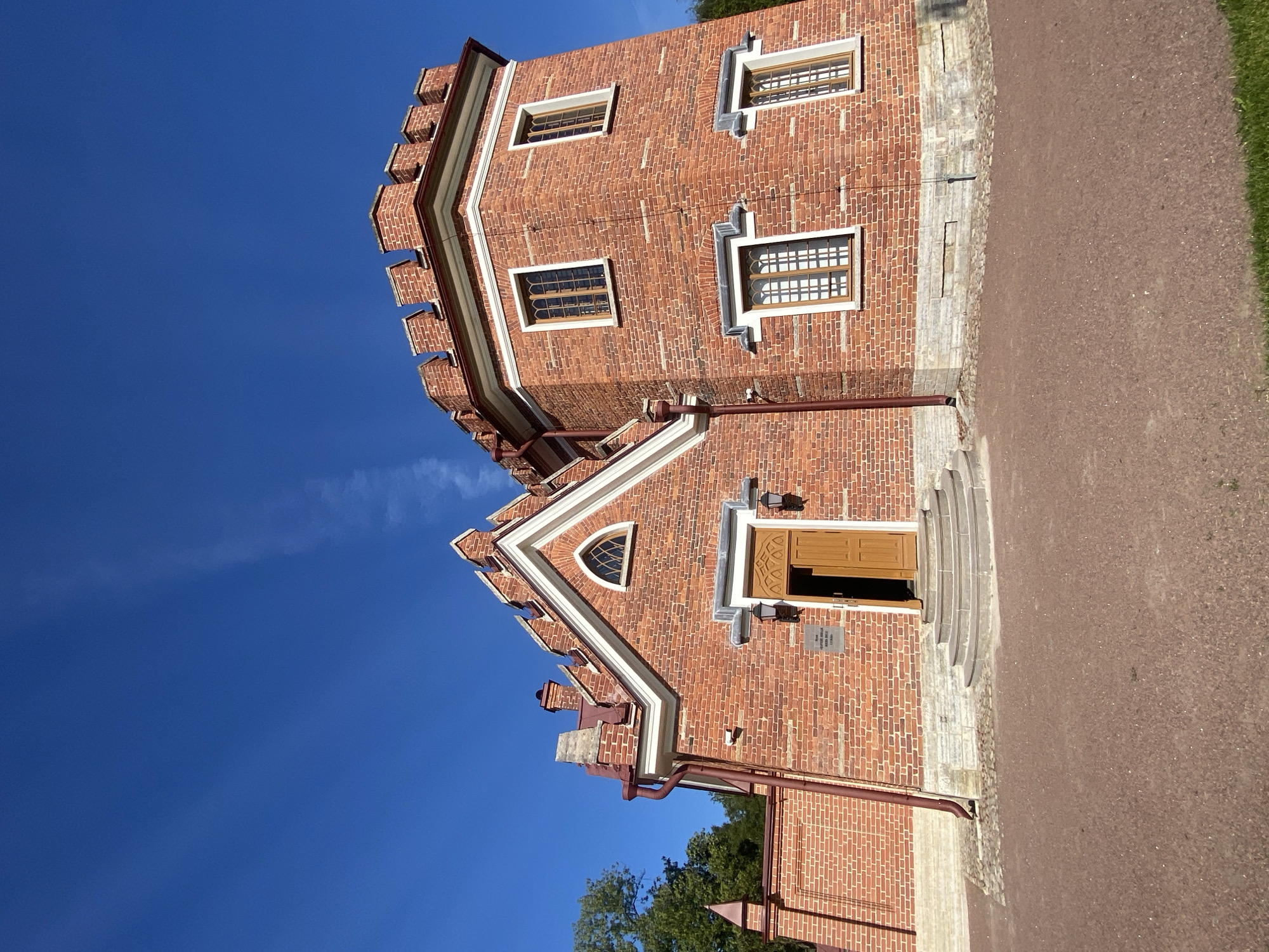 The image shows the Arsenal building in the Catherine Park in Pushkin, Russia, on a clear, sunny day. The building is constructed of red brick with crenellated parapets and arched windows, giving it a castle-like appearance. It features a gabled section with a small, arched window and a larger tower-like section with multiple windows. An open wooden door is visible on the gabled section, with steps leading up to it. Two lanterns flank the doorway. A sign is mounted on the wall above the lanterns. The ground in front of the building is covered with reddish-brown gravel. Green trees and a wall of red brick are visible to the left of the building. The sky is a bright, clear blue with a faint wispy cloud.