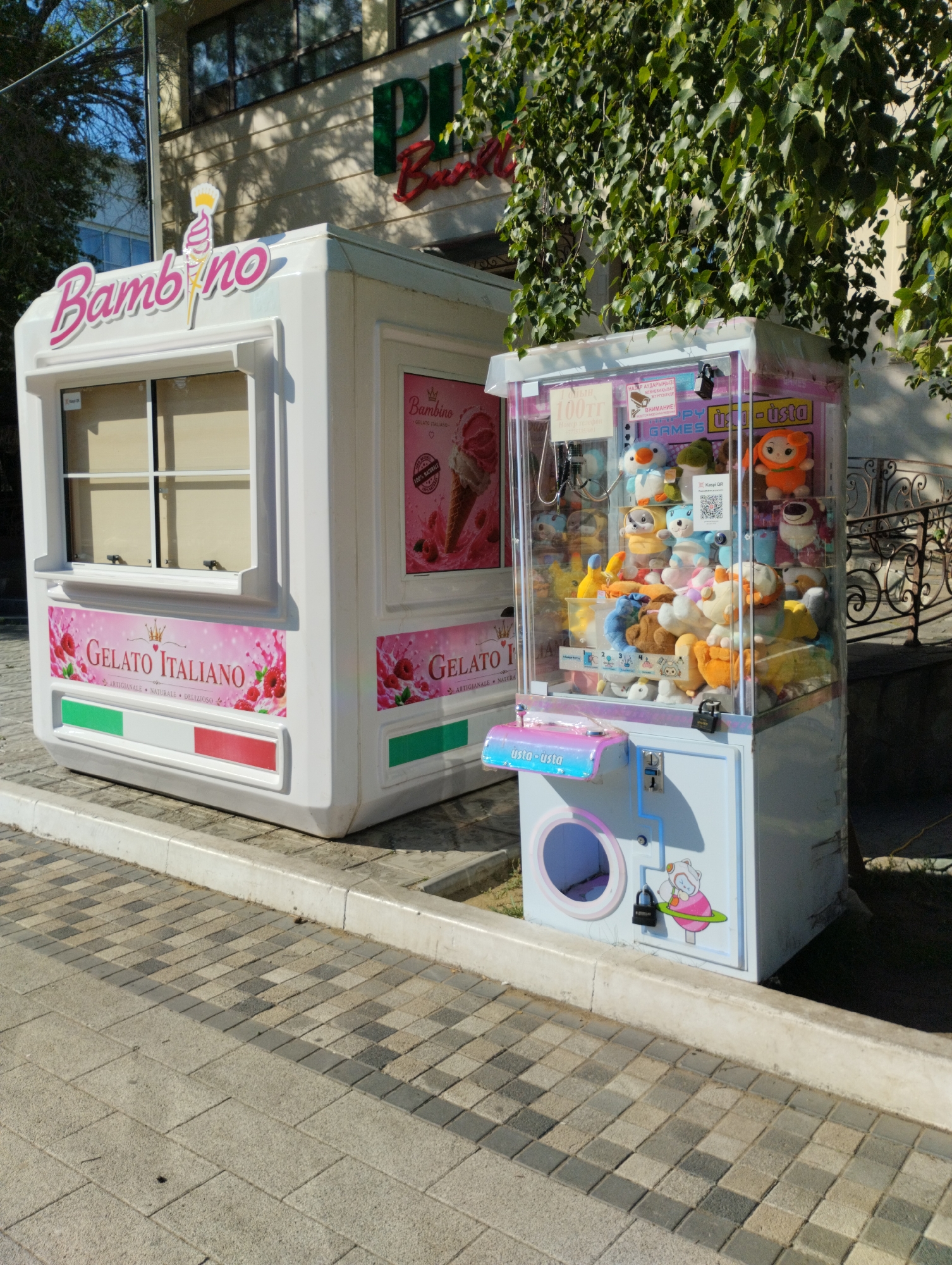 The image captures a street scene in Kostanay, Kazakhstan, focusing on an outdoor ice cream kiosk and a plush toy claw machine. The kiosk, labeled "Bambino" and "Gelato Italiano," is a white, compact structure with a window for service and prominent branding featuring images of ice cream and raspberries. Adjacent to it is a glass-enclosed claw machine filled with various plush toys. The machines are positioned on a paved walkway, with a building and some greenery in the background. The lighting suggests it is daytime, with shadows indicating the sun's position. No people are visible in the immediate vicinity of the machines.
