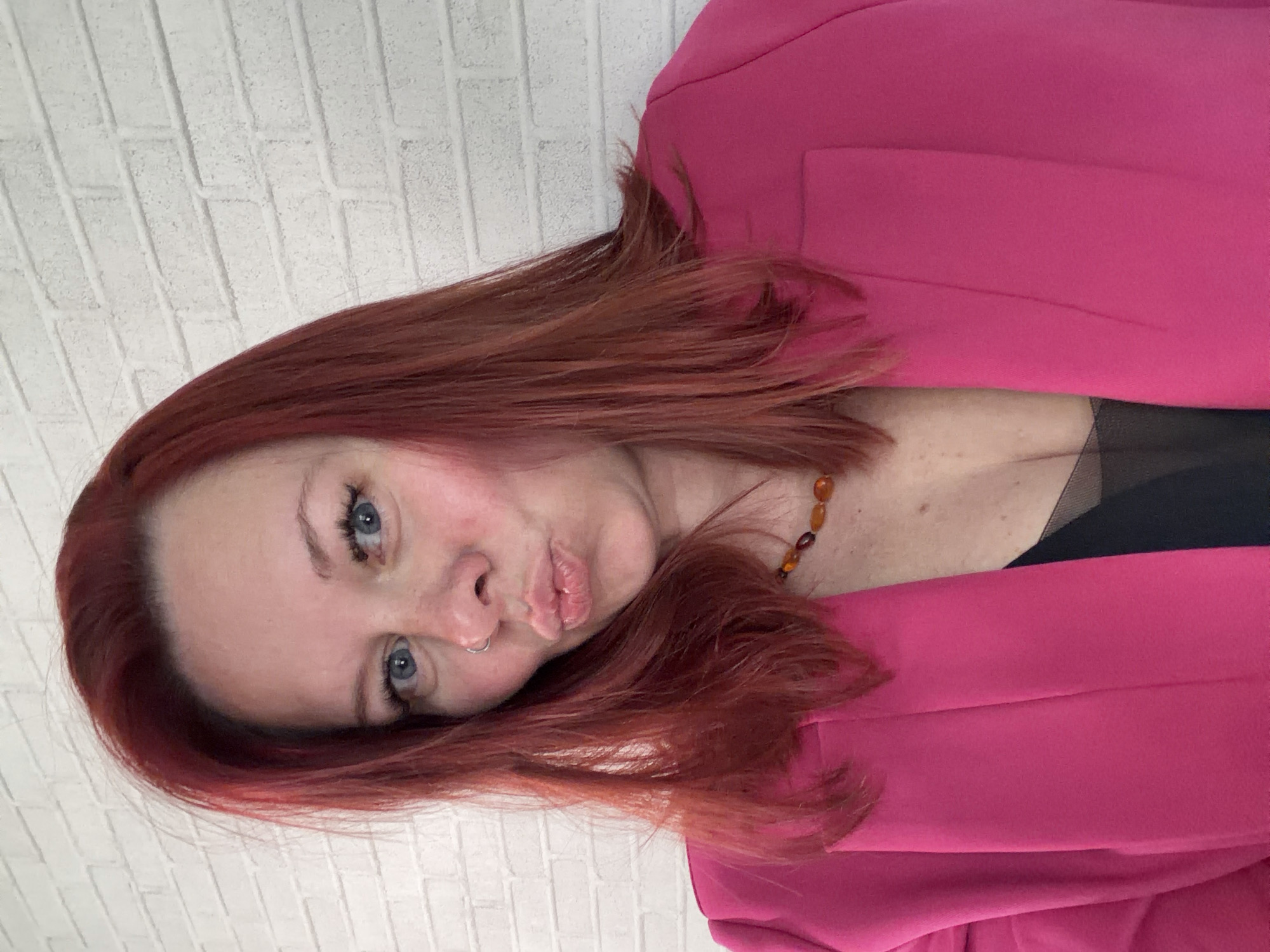 The image shows a woman with red hair and blue eyes wearing a pink blazer and a black top with sheer fabric. She has a nose ring and a necklace with amber-colored beads. The background appears to be a white brick wall. There is no information to indicate this image was taken in Chaikovsky, Russia.
