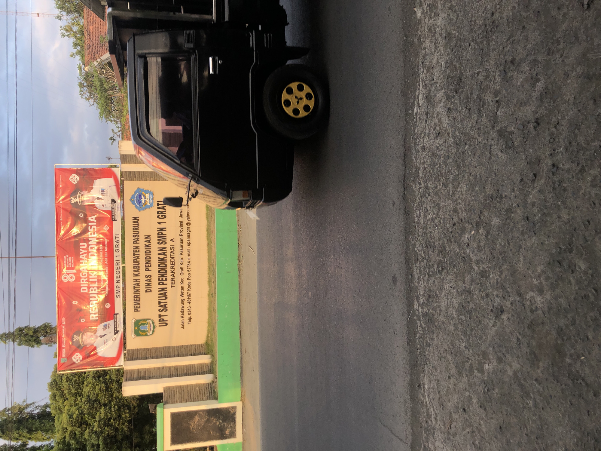 A black pickup truck with yellow wheel accents drives past a wall with signs at SMP NEGERI 1 GRATi in Kedawung Wetan, Indonesia. The wall has a large red banner celebrating "DIRGAHAYU REPUBLIK INDONESIA" with images of officials. Below the banner, a beige sign reads "PEMERINTAH KABUPATEN PASURUAN DINAS PENDIDIKAN UPT SATUAN PENDIDIKAN SMPN 1 GRATi TERAKREDITASI A" followed by an address and contact information for the school. The environment appears to be a street in a semi-urban area with trees and buildings visible in the background. The sky is overcast.
