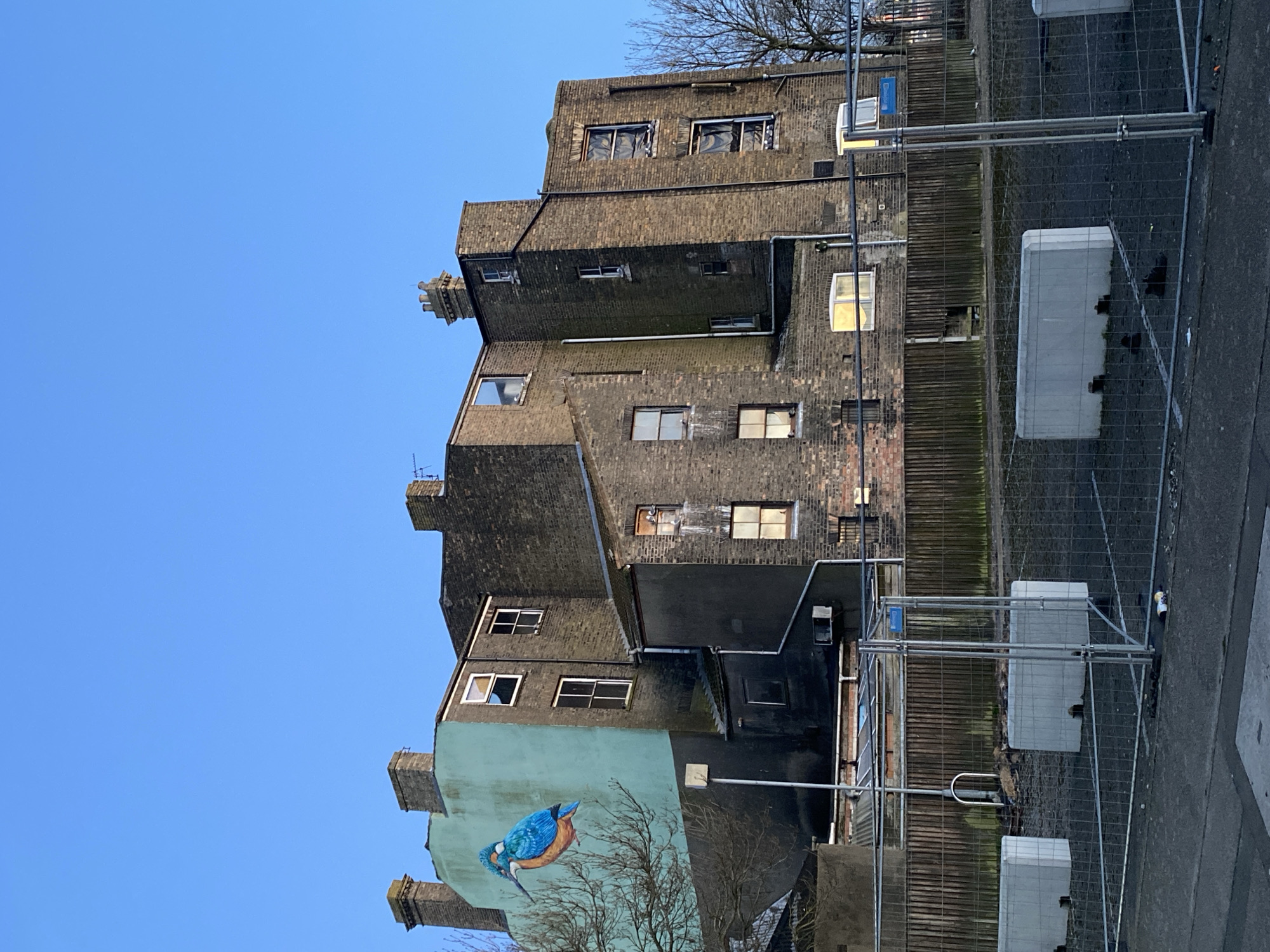 The image captures a multi-story brick building in East Suffolk, United Kingdom. The building appears old, with visible wear and tear. A prominent mural of a blue bird adorns the lower section of the building's facade. A construction fence with white barriers stands in front of the building, suggesting ongoing or recent work. The sky is clear and blue. The image is taken from a ground-level perspective, looking upward at the building. No people are visible in the frame.