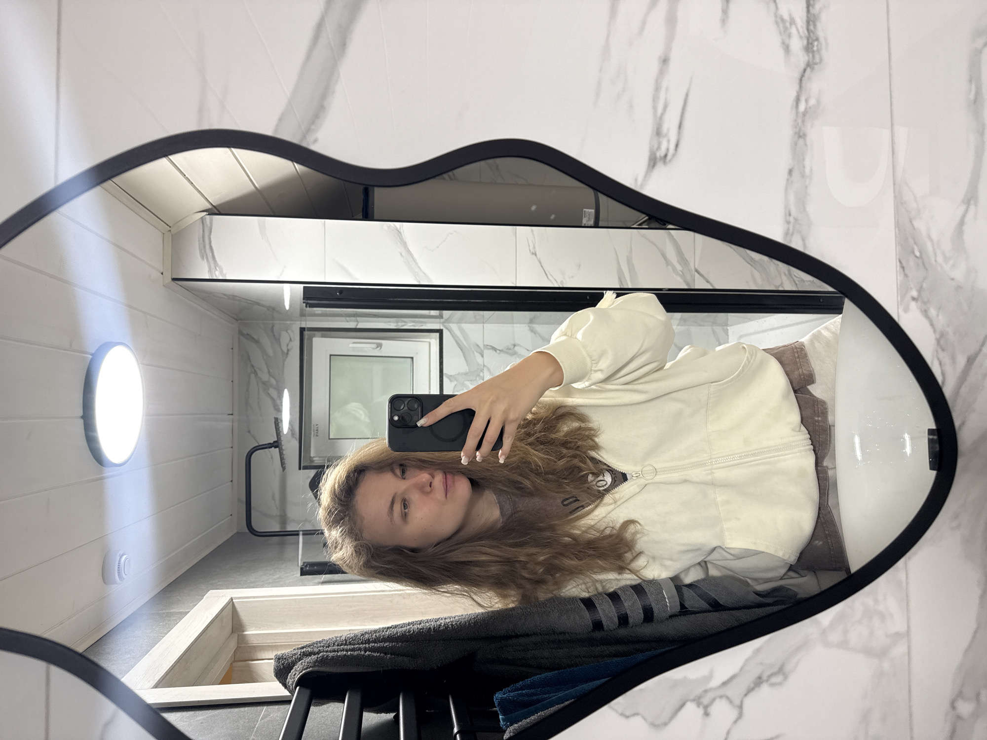 A young woman with long, curly blonde hair takes a selfie in a modern bathroom. She is wearing a cream-colored hooded jacket over a brown t-shirt and appears to be in the process of putting on makeup, with one hand holding her phone and the other near her face. The bathroom features marble-patterned tiles on the walls and a white sink. A black towel rack with towels hangs on the wall to the left. The scene is well-lit, possibly by overhead lighting visible in the reflection on the ceiling. The image does not contain any information that places it in Prokudino, Russia.