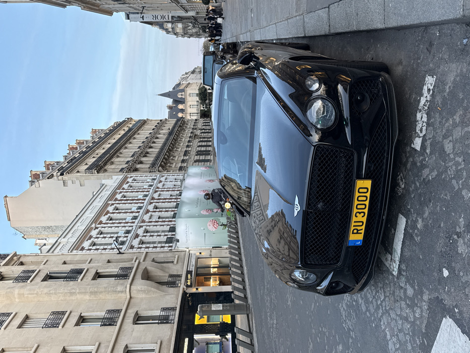 A black Bentley Continental GT coupe is parked on the right side of a cobblestone street in Paris, France, specifically identified as Rue de la Paix. The vehicle bears a yellow Luxembourgish license plate, "L RU 3000." Its glossy surface reflects the surrounding urban architecture and the sky.
To the left of the Bentley, a person wearing a dark helmet and jacket is operating a moped, moving away from the viewer in the street. The street surface consists of dark asphalt interspersed with cobblestones.
On the left side of the street, a line of light-colored bollards separates the sidewalk from the roadway. Behind these bollards, commercial establishments are visible: a "Dinh Van" jewelry store with illuminated displays, adjacent to a construction hoarding featuring "Van Cleef & Arpels" branding and pink floral motifs. Beyond these, multi-story Haussmannian buildings with classic Parisian balconies extend down the street.
On the right side of the street, further down, a group of approximately eight pedestrians is observed on the sidewalk near a building displaying a "DIOR" sign. Another dark vehicle, likely an SUV, is parked further along the curb behind the Bentley.
The background reveals a continuous streetscape of ornate Parisian buildings under a clear, light blue sky. A distinctive spire or turret is visible on a building further down the street, providing an architectural landmark. The scene is illuminated by natural light, consistent with morning or late afternoon.