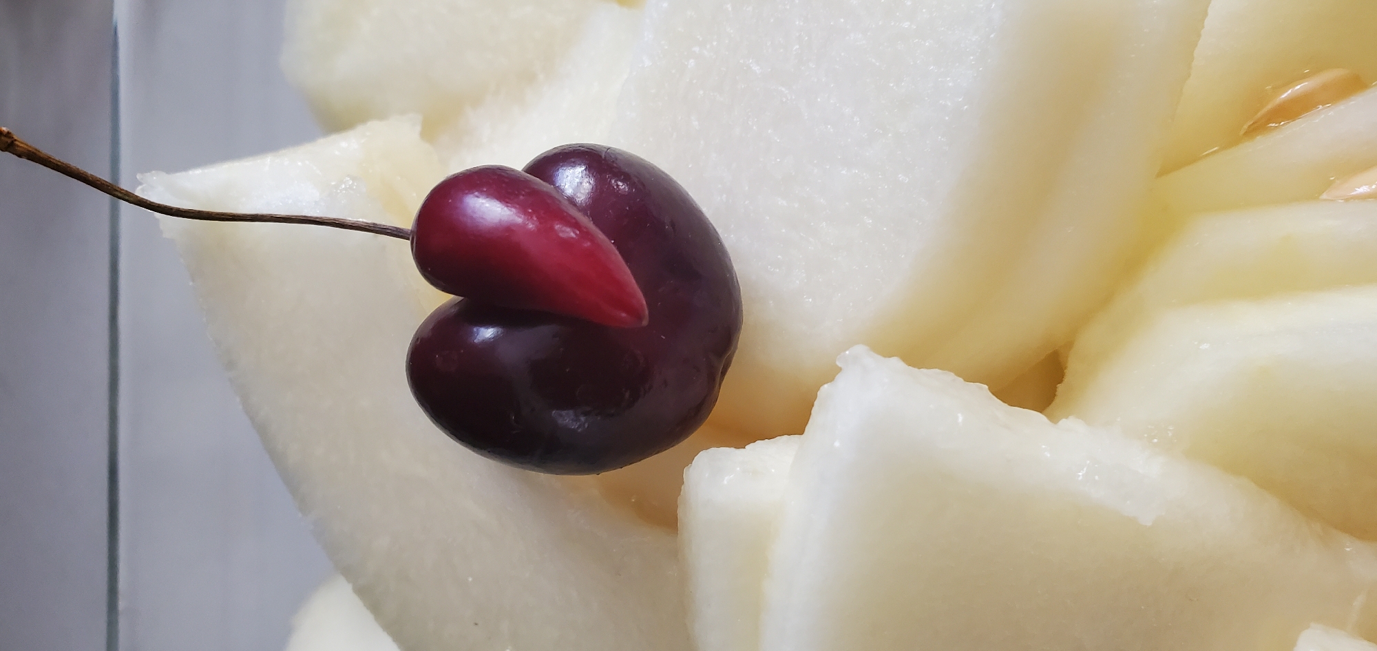 A close-up, vertical shot features a cluster of pale, sliced fruit, likely melon, with a single dark red cherry perched on top. The cherry is unusual in appearance, appearing to be two conjoined cherries or a cherry with an additional growth. It is positioned slightly off-center towards the right of the frame, with its stem extending upwards and out of view. The fruit slices are arranged in a layered fashion, creating a textured background. The lighting is bright, highlighting the smooth surfaces of the fruit. The scene is a still life composition, focusing on the textures and colors of the fruit. No specific location in Νέα Σάντα, Greece is discernible from the image.