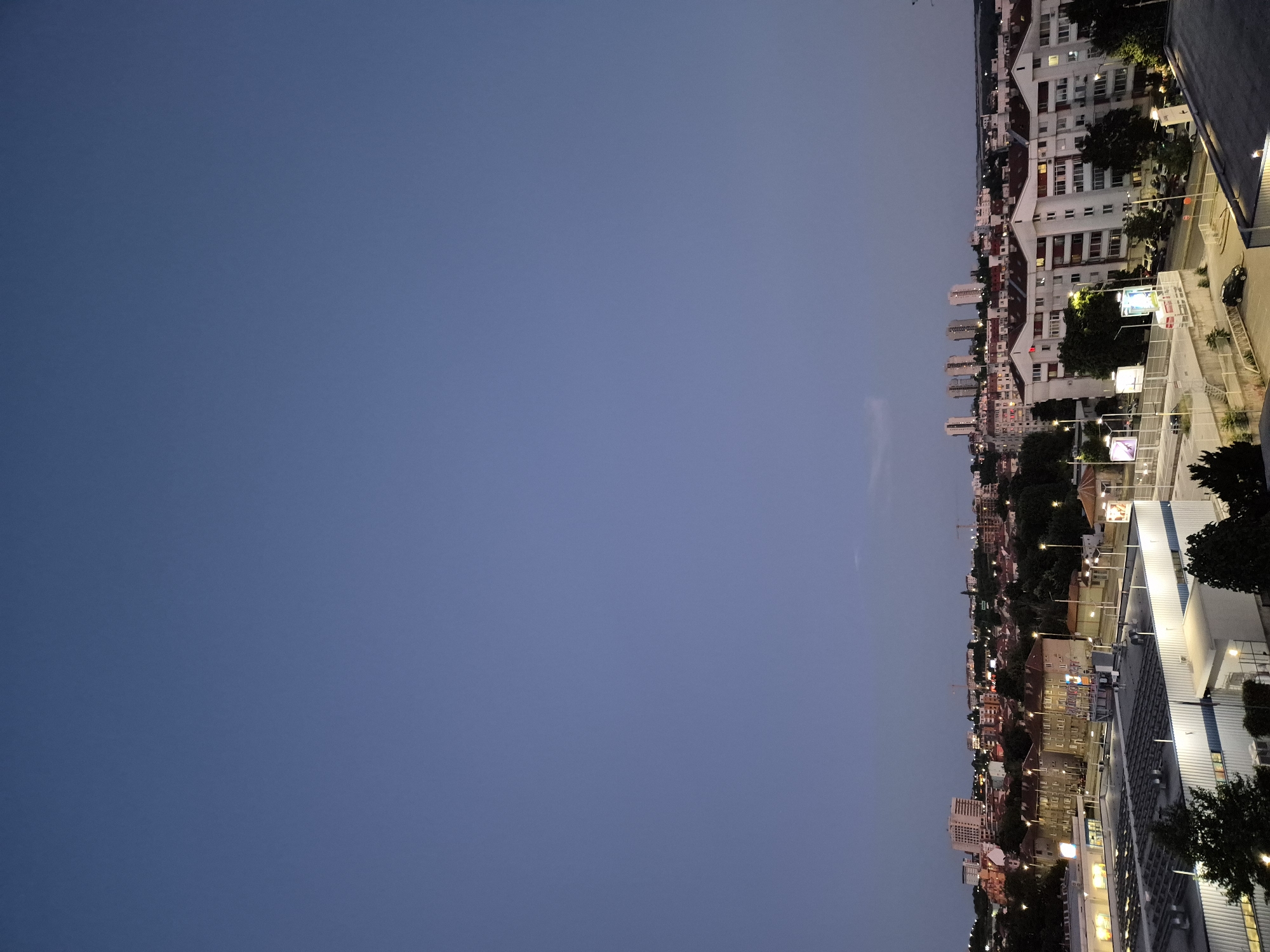 This image shows an aerial view of a city at dusk. The sky is a deep blue, and the city lights are beginning to twinkle. In the foreground, there are several large, modern buildings, likely commercial or industrial, with bright lighting. Beyond these, a dense urban landscape unfolds with a mix of residential buildings, featuring red-tiled roofs, and taller apartment blocks. Trees are interspersed throughout the city, adding patches of dark green. Streetlights are visible, illuminating roads and walkways. In the distance, the city skyline is punctuated by more apartment buildings and some construction cranes, suggesting ongoing development. The overall impression is of a bustling urban area transitioning from day to night.