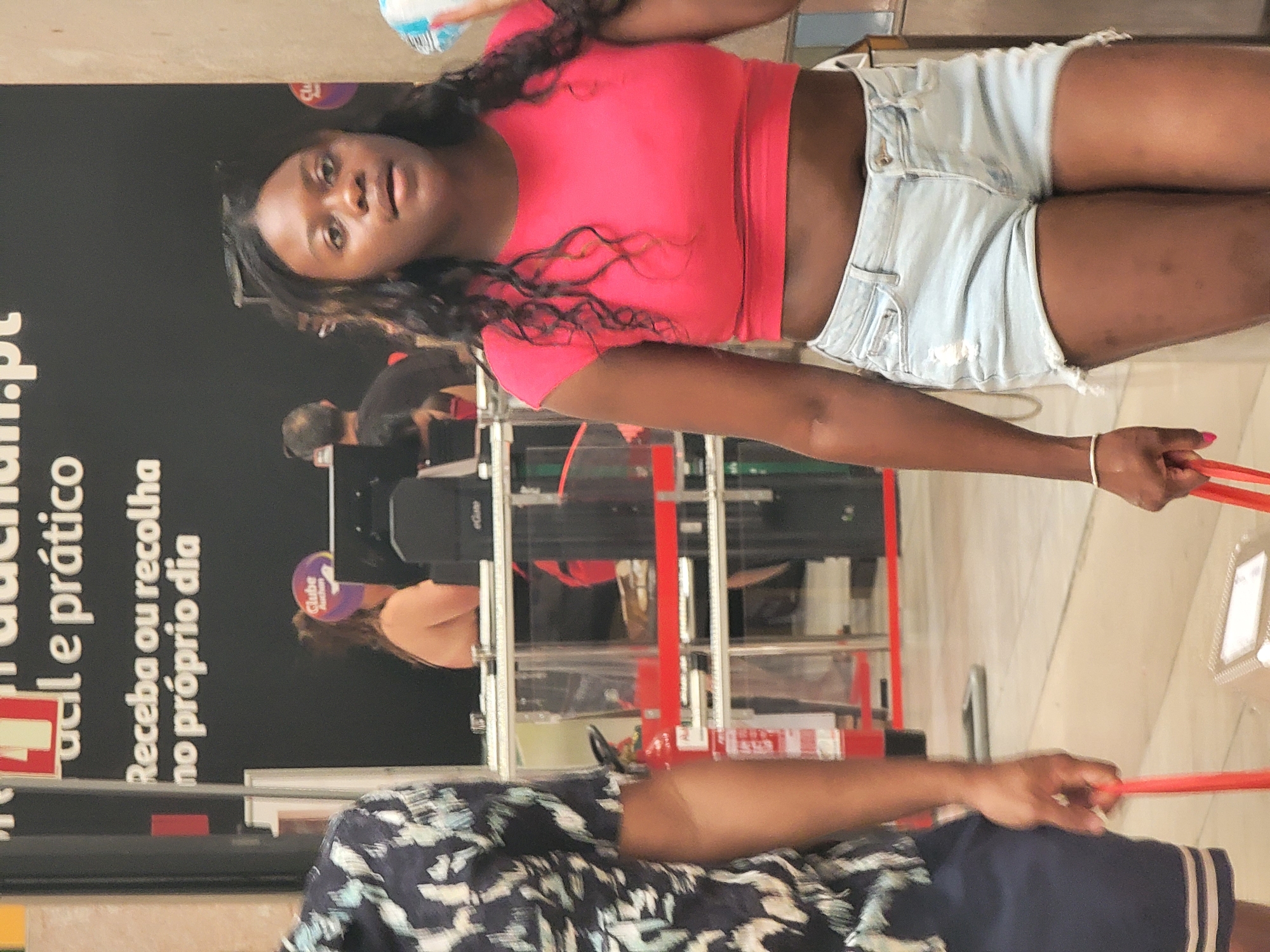 The image shows two people in what appears to be a retail store. In the foreground, a person on the left is wearing a dark blue and white patterned shirt and navy shorts, carrying a red bag handle. To the right, a young Black woman is wearing a pink crop top and light blue denim shorts, also carrying a red bag handle. She is looking towards the right of the frame. In the background, there is signage with Portuguese text, indicating a location in Faro, Portugal. The text on the signage reads "Receba ou recolha no próprio dia," which translates to "Receive or collect on the same day." Further in the background, there are other individuals, checkout counters, and retail displays. The environment appears to be indoors, possibly a supermarket or a similar establishment.