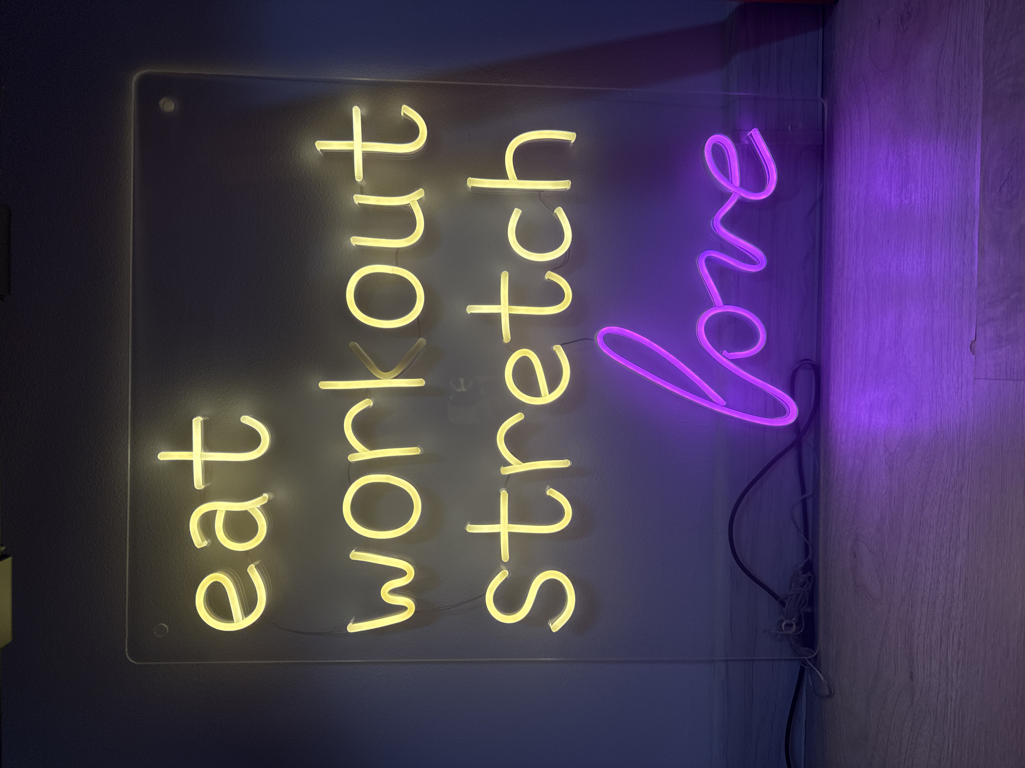 The image depicts a neon sign with the words "eat workout stretch love." The words "eat," "workout," and "stretch" are illuminated in a yellow neon color, while the word "love" is illuminated in purple. The sign appears to be mounted on a wall, with a light-colored wall on the left and a light-colored wooden panel on the right. The image does not contain any information that would allow one to determine the location of the image in Tyumen, Russia.