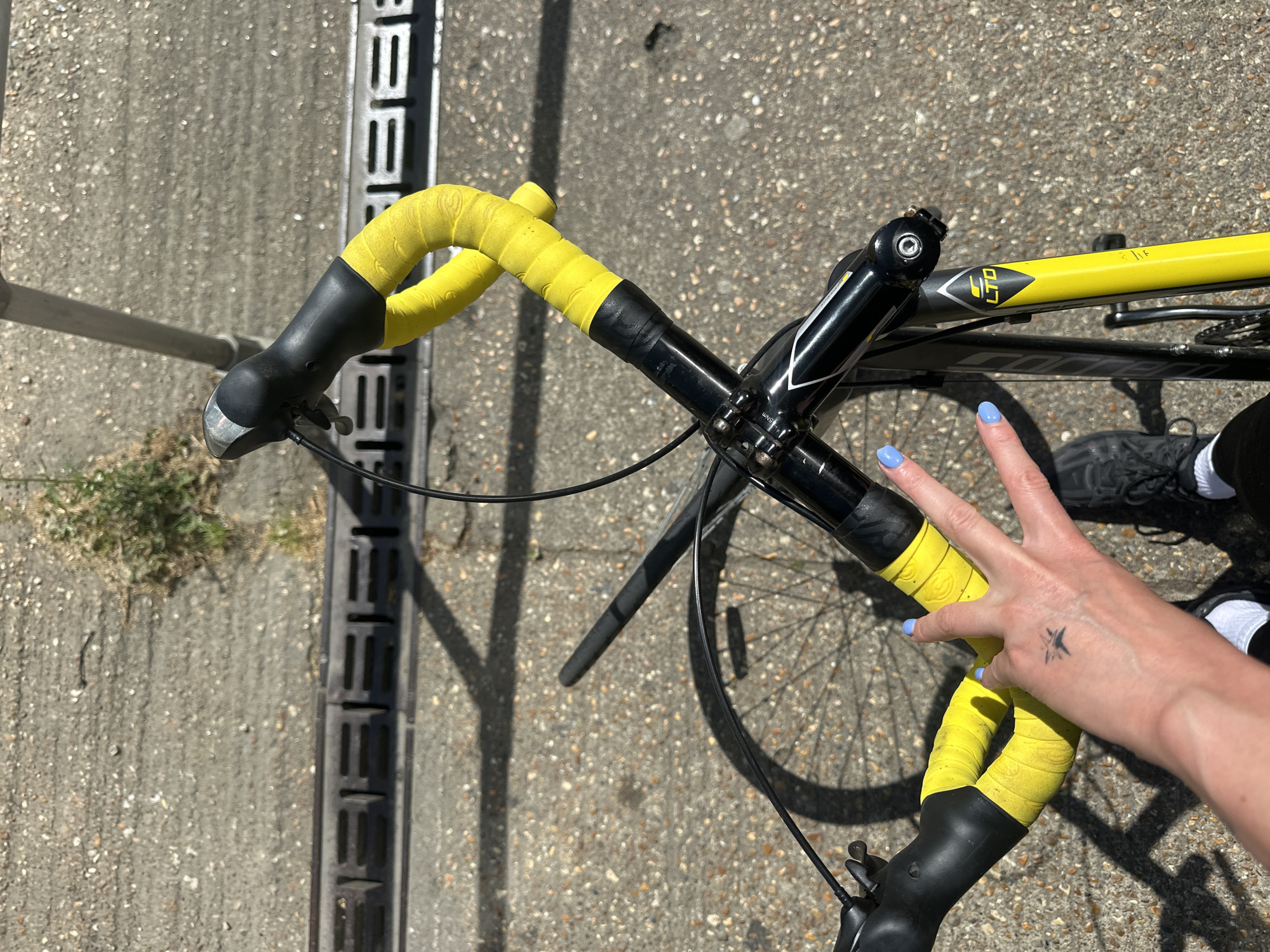 Overhead shot of a bicycle with yellow handlebar tape on a grey concrete surface, possibly a street in Tunbridge Wells, United Kingdom. The bike's frame is black and yellow, with "LTD" visible on the frame. A hand with blue nail polish and a tattoo of a bird is giving a peace sign over the handlebars. A black shoe and white sock are visible in the background. A metal grate runs parallel to the bike, with some vegetation nearby. The lighting is bright, suggesting a sunny day.