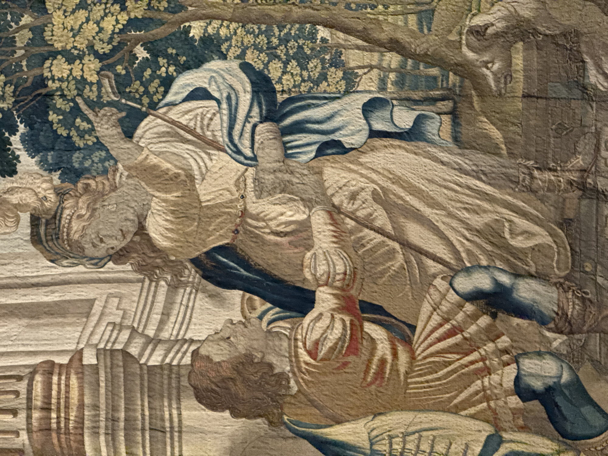 This tapestry depicts a scene with multiple figures in what appears to be an outdoor setting, possibly a garden or forest, suggested by the lush foliage and trees in the upper left. Two prominent figures, a woman and a man, are central to the composition. The woman, in the upper part of the image, is dressed in flowing garments of white and blue and holds a staff or rod. Her head is turned, and her expression is difficult to discern clearly due to the tapestry's texture and detail. She seems to be in motion, perhaps gesturing or walking.
Below and to the right of the woman lies a man, also richly dressed in attire that includes striped fabric, red accents, and blue leg coverings. He appears to be either lying down or wounded, with his head turned to the side. His posture suggests a state of repose or vulnerability. In the lower left, architectural elements resembling steps or platforms are visible, rendered in muted tones. The overall color palette is dominated by earthy browns, blues, greens, and creams, typical of historical tapestries. The lighting seems subdued, consistent with an indoor or shaded outdoor environment. There is no visible text in the image. The scene evokes a narrative, possibly from mythology, history, or a religious story, though the specific event or context is not explicitly clear from the visual information alone. The style suggests a Renaissance or Baroque period of art. The location context of Santiago de Compostela, Spain, might suggest a connection to a pilgrimage or a historical event relevant to the region, but this is not directly represented in the tapestry itself.