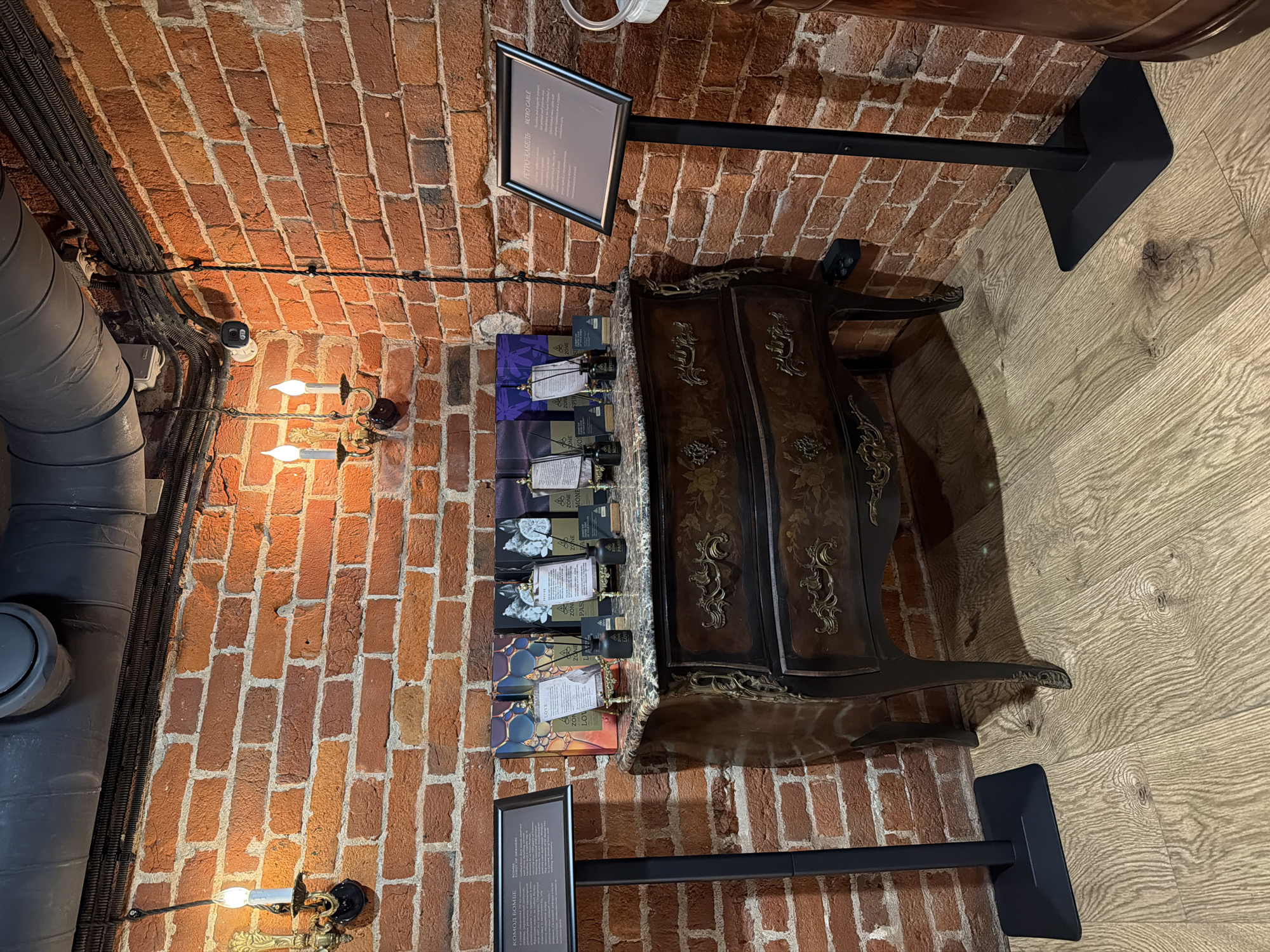 The image shows an interior space with a brick wall and wooden floor. A large, dark-colored pipe runs vertically along the left side of the brick wall. On the wall, there are two sconces with candle-like lights.
A dark wooden dresser with decorative details stands against the brick wall, along with a display rack with several packages of goods. There are two signs on stands with written material.
Based on the "RETRO CABLE" writing and the style of products displayed, this is likely a commercial space of some kind. Given the Russian text on the signs, it's reasonable to assume this image was taken in Russia. Since the text on the sign appears to be in Cyrillic, the city could be Moscow.