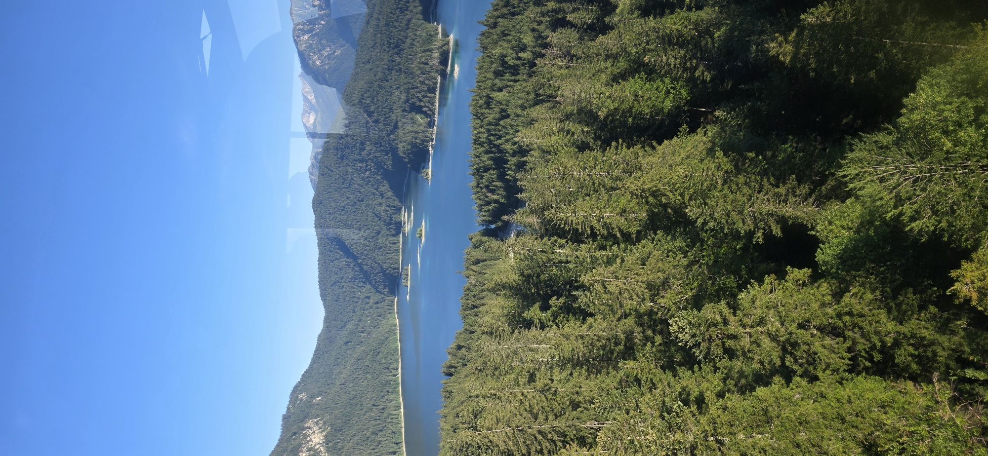 This image depicts a panoramic view of the Eibsee, a picturesque lake located at the foot of Zugspitze mountain in Grainau, Germany. The scene is dominated by the crystal-clear blue waters of the lake, which is dotted with several small, tree-covered islands. Surrounding the lake are lush, dense green forests that climb the slopes of the majestic mountains. The sky above is a brilliant, cloudless blue, indicating a clear and sunny day. Reflections of the sky and surrounding landscape are visible on the surface of the lake. The perspective suggests the photograph was taken from an elevated position, possibly a cable car or a high vantage point, looking down and across the lake and the surrounding alpine terrain. The image conveys a sense of natural beauty and tranquility.