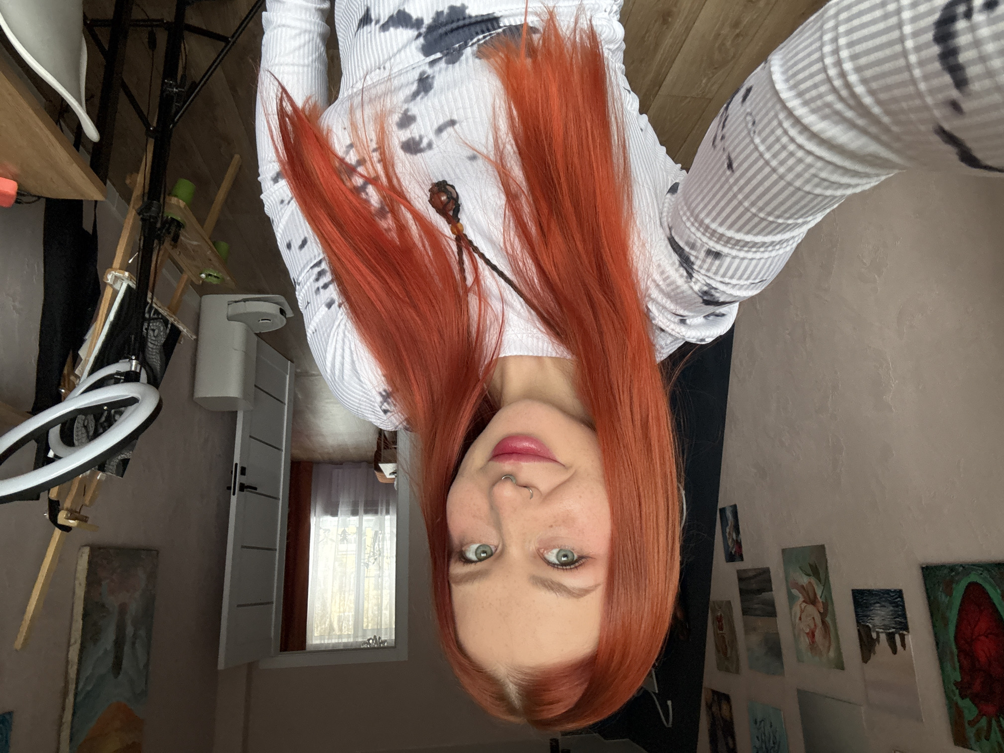 This image, taken in Rostov-on-Don, Russia, depicts a female subject in an indoor residential setting, appearing to take a selfie. The subject, positioned centrally, has long, straight, vibrant red hair, fair skin with freckles, light-colored eyes, and a silver septum piercing. She is wearing pink lipstick and has a subtle smile. Her attire consists of a long-sleeved top with a white ribbed texture on the sleeves and a tie-dye pattern of black and grey on the torso. A dark braided necklace with a reddish stone pendant is visible around her neck.
The background reveals a room with light-colored, possibly textured walls. On the left wall, numerous small to medium-sized artworks are displayed, including a painting of a red anatomical heart on a dark green background, landscapes (one with sea stacks, one with water), a floral piece, and abstract art. To the right, an open white door with horizontal panels and a dark handle leads to another room. Through the doorway, a window with sheer white and thicker orange curtains is visible, allowing natural light to enter.
Near the right edge of the frame, a wooden art easel holds a canvas. In front of the easel, a black tripod stands, equipped with a white ring light. A light-colored wooden desk or table and a white chair are partially visible on the far right. A white cylindrical object, possibly a robotic vacuum cleaner or air purifier, rests on the light brown wood or laminate floor near the door. Another painting with blue and yellow tones hangs on the wall above the easel. The overall environment suggests an artist's personal space or home studio.
