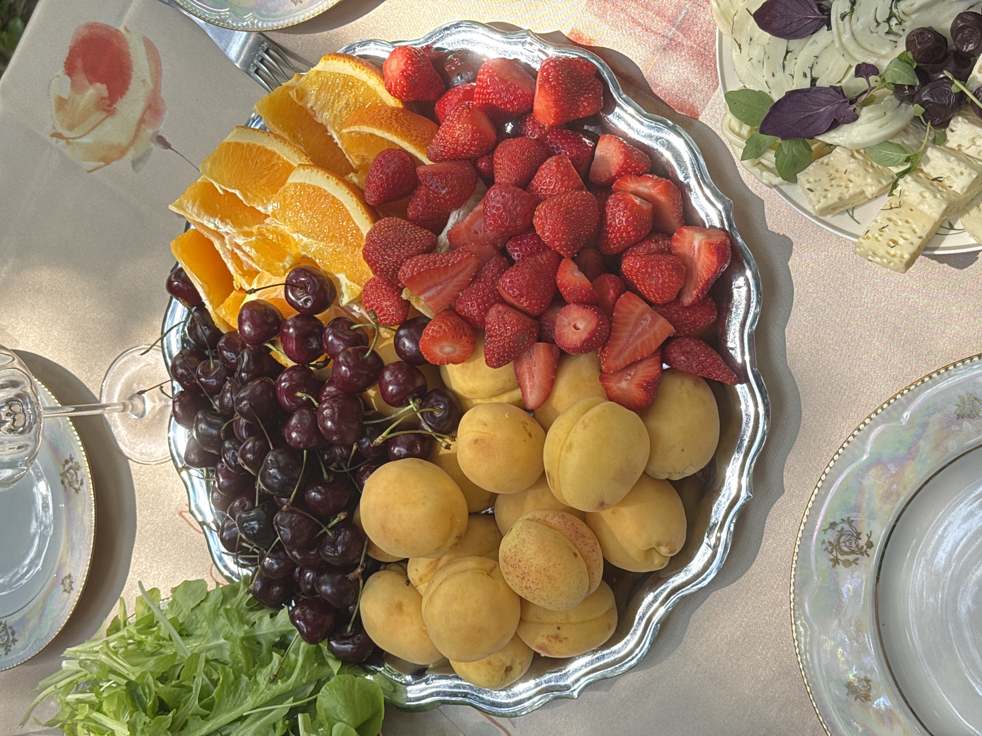 The image shows a close-up, high-angle view of a table set for a meal, likely outdoors. A silver platter filled with a vibrant assortment of fresh fruit is the main subject. The platter contains dark red cherries, bright orange orange slices, whole yellow apricots, and halved strawberries. To the left of the platter, a bunch of green leafy vegetables, possibly arugula or a similar salad green, is visible. In the foreground and to the right, another plate holds a collection of white cheeses, some cubed and some thinly sliced and rolled, garnished with fresh green herbs and a few dark purple basil leaves. The table is covered with a light-colored tablecloth featuring a subtle floral pattern. In the background, partially visible, are decorative plates and a stemmed glass. The lighting suggests it is daytime, with shadows indicating a bright, possibly sunny, environment. The scene appears to be set in a domestic or garden setting, consistent with a meal in Козино, Russia, during favorable weather conditions. There are no people or animals visible in the image.