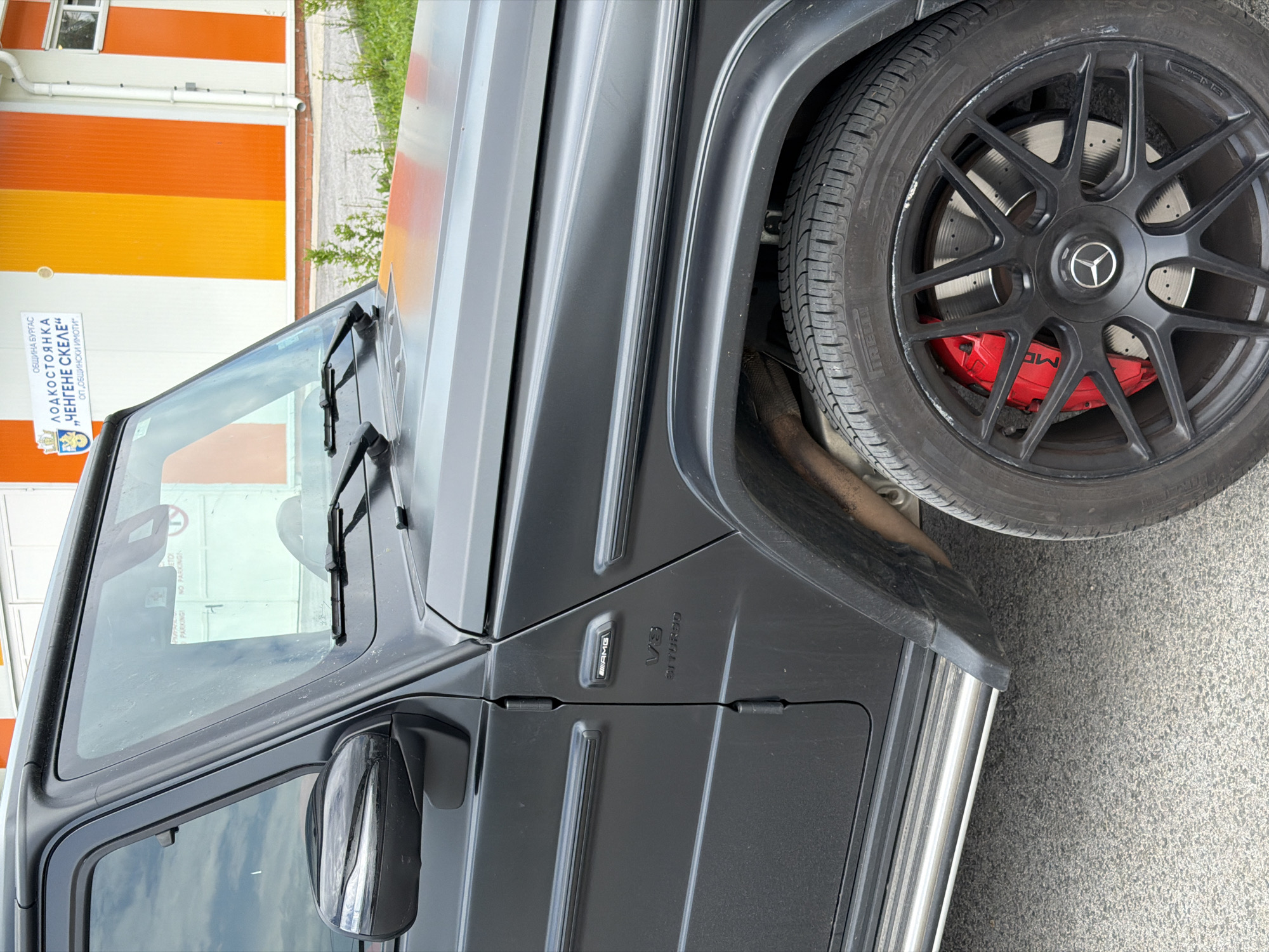 The image shows the front end of a matte black Mercedes-Benz G-Class (G-Wagon) AMG, oriented at a slight angle. Visible features include the front wheel with black rims and red brake calipers, the front fender, windshield, side mirror, and the "V8 BITURBO" emblem. A sign in Cyrillic is visible in the background, partially obscured by the car's windshield. The sign reads "ОБЩИНА БУРГАС ЛОДКОСТОЯНКА „ЧЕНГЕНЕ СКЕЛЕ“ ОП, ОБЩИНСКИ ИМОТИ". This indicates a location in the city of Burgas, Bulgaria, specifically at or near the "Chengene Skele" boat parking area. The building in the background appears to be orange and white. The setting is outdoors, likely a parking lot or a street. The weather is daytime, with fair lighting.