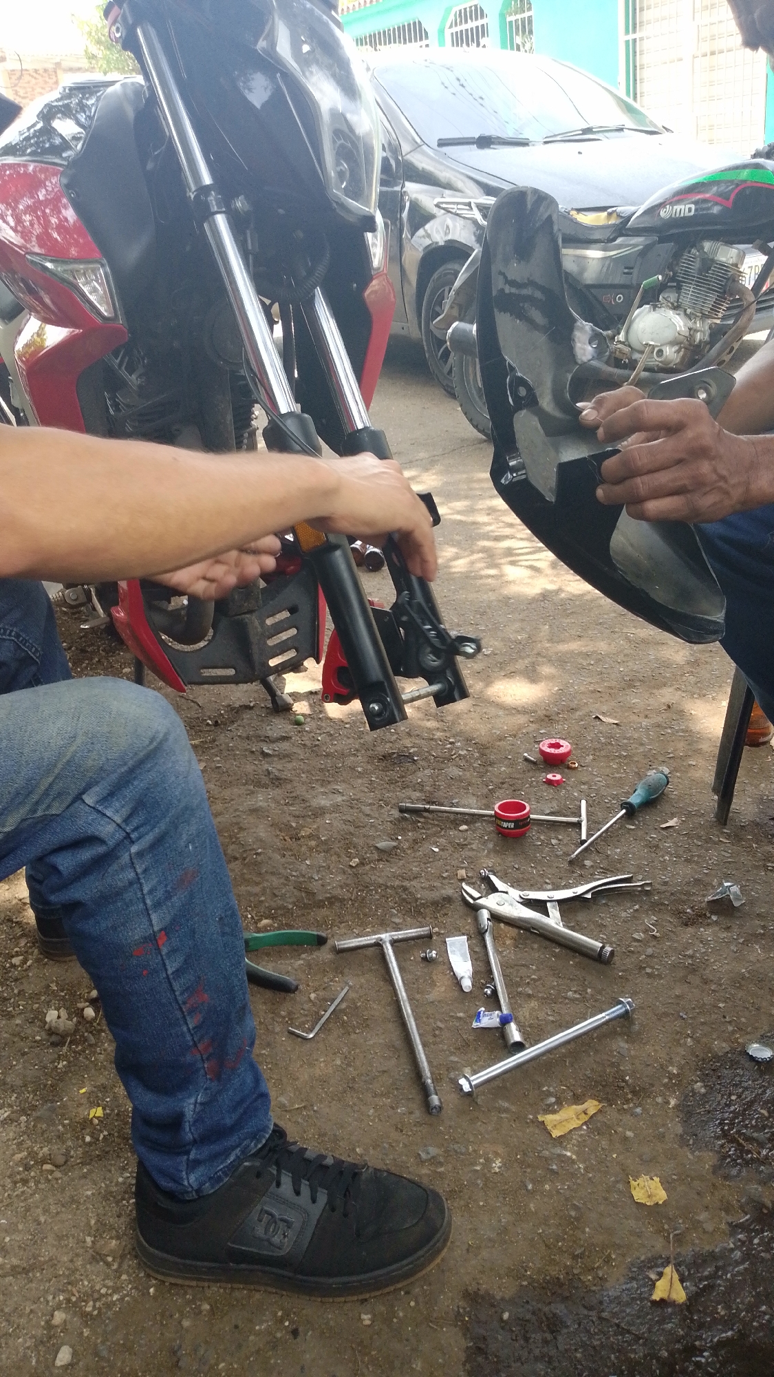 The image depicts a motorcycle repair scene in what appears to be Tinaquillo, Venezuela. Two men are actively working on the front suspension of a red motorcycle. One man is holding a disassembled fork, while the other is working with a black motorcycle panel. Tools such as wrenches, pliers, screwdrivers, and tubes of lubricant are scattered on the ground around them. One of the men is wearing jeans and black DC shoes. In the background, there is a black car parked and another motorcycle. The ground is dirt. A blue and white building is visible in the background.