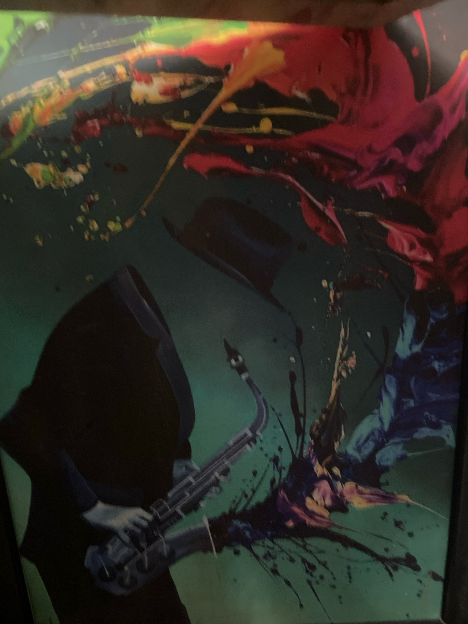 The image is a close-up of a painting or digital artwork featuring abstract splashes of vibrant colors, including red, pink, yellow, green, and blue, against a dark teal background. In the foreground, a dark silhouette of a person wearing a hooded cloak and a hat is depicted playing a saxophone. The saxophone itself is rendered in a muted, metallic tone, with intricate details visible on its keys and body. The abstract paint splatters appear to emanate from or surround the figure and the instrument, suggesting a dynamic and energetic representation of music. The overall impression is one of artistic expression and movement, with the artist's style being bold and somewhat chaotic. There is no discernible information within the image to suggest a specific location in the city of Abuja, Nigeria.