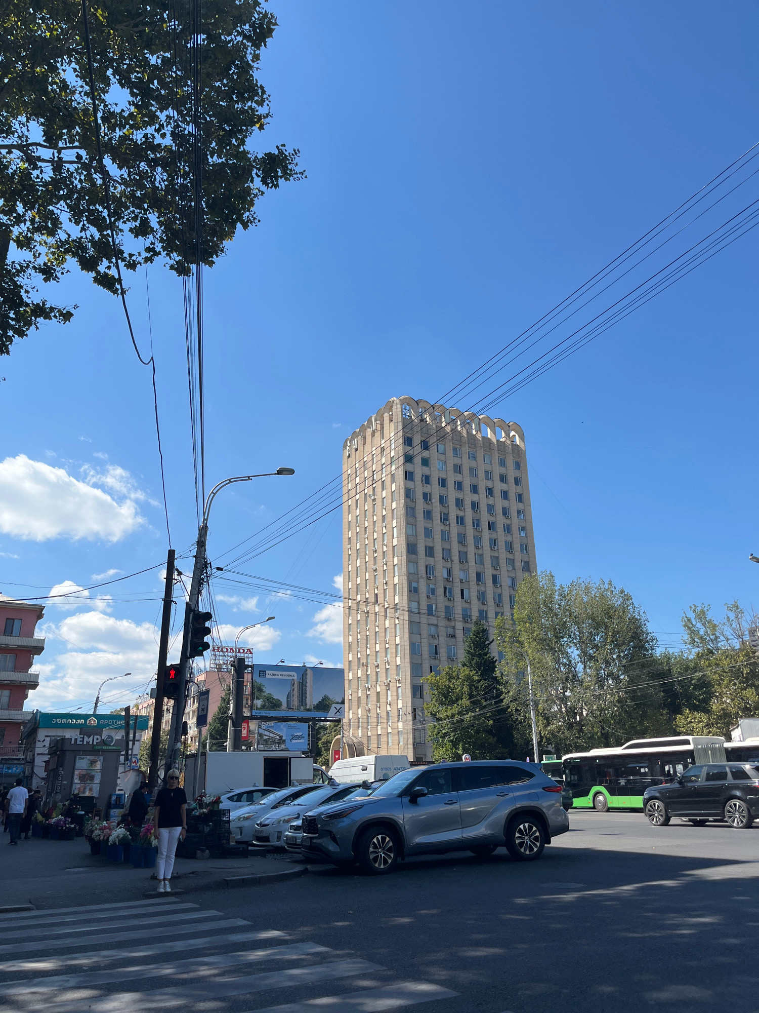 In Tbilisi, Georgia, on a sunny day, a street scene unfolds with a mix of urban elements. A tall, beige building with arched decorative features on its roof dominates the skyline, flanked by trees. In the foreground, a pedestrian crossing is visible on the asphalt road, with several cars parked and in motion, including a silver SUV and a black SUV. A green bus is also present on the road. A traffic light shows a red signal. To the left, a building with pink balconies and a flower stall are situated near a street light and utility poles with numerous wires. A woman in white pants and a dark shirt stands on the pedestrian crossing. Several people are walking on the sidewalk. Billboards with advertisements are mounted on buildings in the background. The sky is clear blue with scattered white clouds.