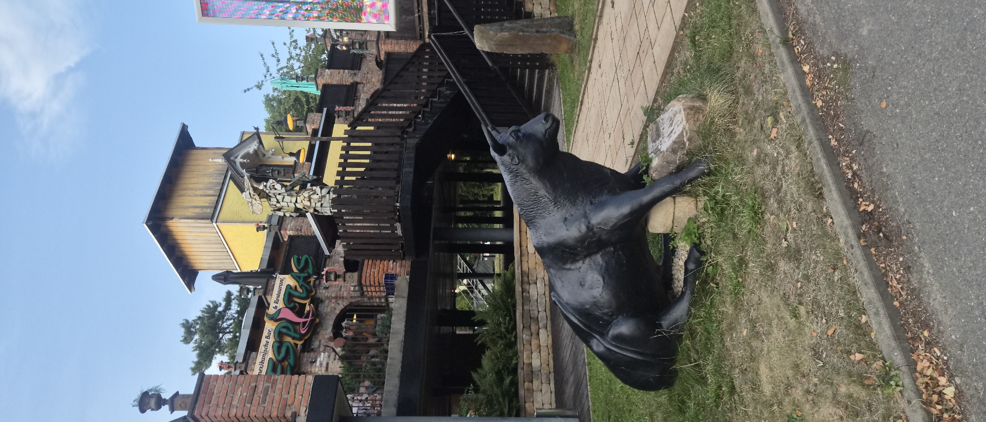 The image shows a dark, black statue of a bull in a seated position on grass in front of a Mexican restaurant named "ESPITAS" in Chemnitz, Germany. The bull statue is large and appears to be made of metal, with blue eyes. The restaurant building is multi-story with a yellow upper section and brick lower sections. A wooden staircase leads up to the restaurant. The sky is blue with some clouds. There are trees and other greenery visible in the background. The scene appears to be a street view with a sidewalk and road in the foreground.