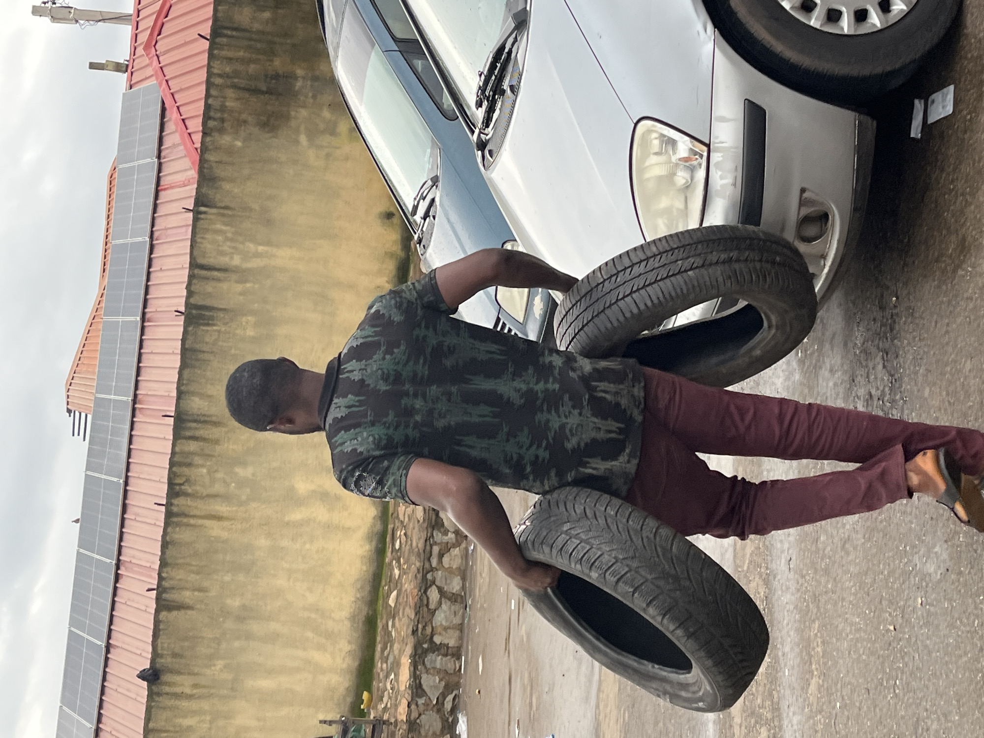 A Black male is walking from right to left, carrying two tires. He is wearing a dark green shirt with a tree-like pattern and maroon pants, with brown sandals on his feet. He is in what appears to be a street or a parking lot, with two cars parked next to him. The car to his left is a light blue sedan, and the car to his right is a silver sedan. In the background, there is a building with a red roof and solar panels. The ground is wet and appears to be made of asphalt. The overall lighting suggests an overcast day. The location is identified as Bukuru, Nigeria.