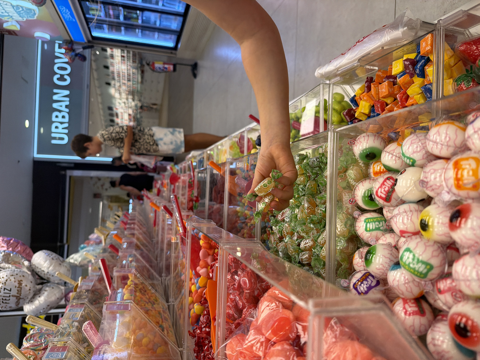 The image depicts a candy aisle in a store, likely a supermarket or a specialized candy shop, in Aldaia, Spain. The foreground is dominated by rows of clear plastic containers filled with various types of candies. In the center foreground, a person's hand is reaching into a bin of individually wrapped, green and yellow candies. To the left, large bins contain red-wrapped candies and pink, round candies. Further back and to the right, a bin is filled with eye-shaped candies, some with green irises and others with blue. Next to these, a bin contains brightly colored, individually wrapped cube-shaped candies in shades of orange, yellow, blue, and red.
In the background, a young boy with short brown hair and wearing a patterned t-shirt and tie-dye shorts is standing and looking towards the candy bins. He appears to be a customer. Behind him, a sign with illuminated blue letters reads "URBAN COVE". Other shoppers are visible in the blurred background, as are shelves stocked with products. The overall environment suggests an indoor commercial setting with bright lighting. Balloons, possibly for a celebration, are visible in the upper left portion of the image.