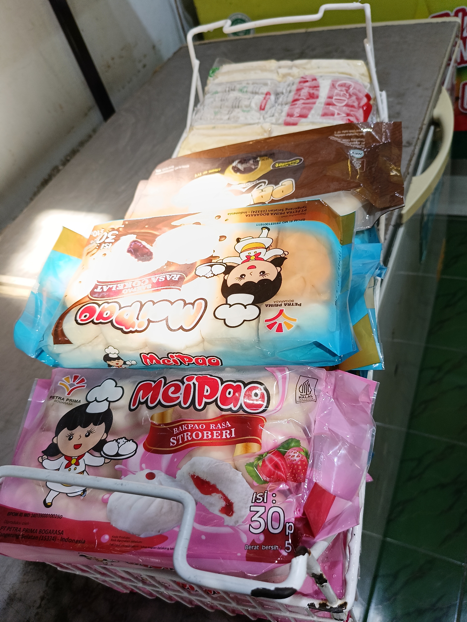 A rack of frozen bao buns is displayed in what appears to be a small grocery store or market stall in Manisrenggo, Indonesia. The buns are packaged in colorful plastic wrappers, with labels indicating flavors such as chocolate and strawberry. The lighting suggests an indoor setting, possibly a refrigerated display case. The background is blurred, but a glimpse of a concrete wall and a green tiled floor is visible.
