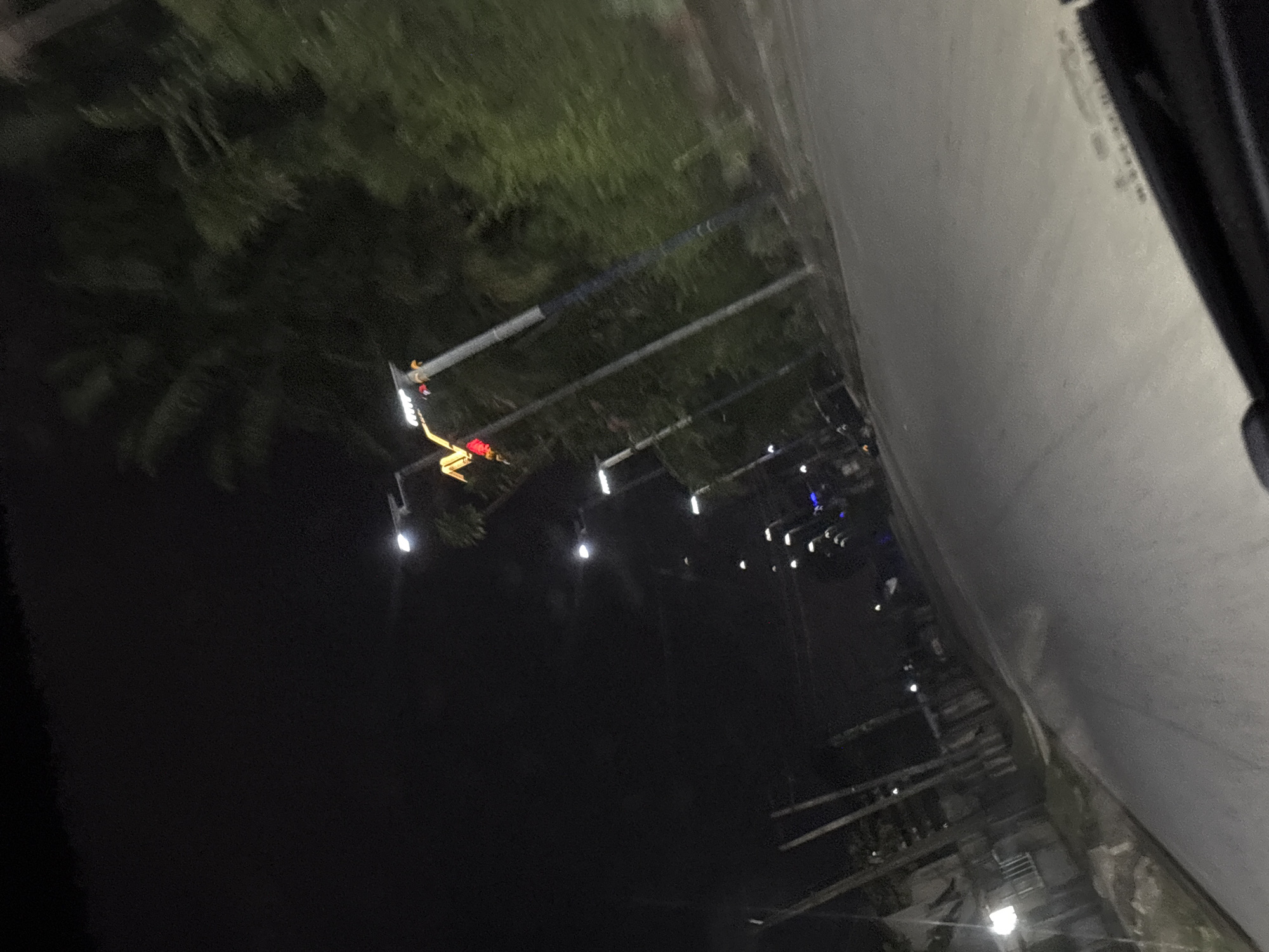 The image shows a dark street at night in Port Harcourt, Nigeria, illuminated by streetlights. There are trees and foliage visible on the left side of the frame. Several streetlights are present, suspended by metal poles. A section of what appears to be a concrete structure or building is visible on the right side of the image. There are no people visible.